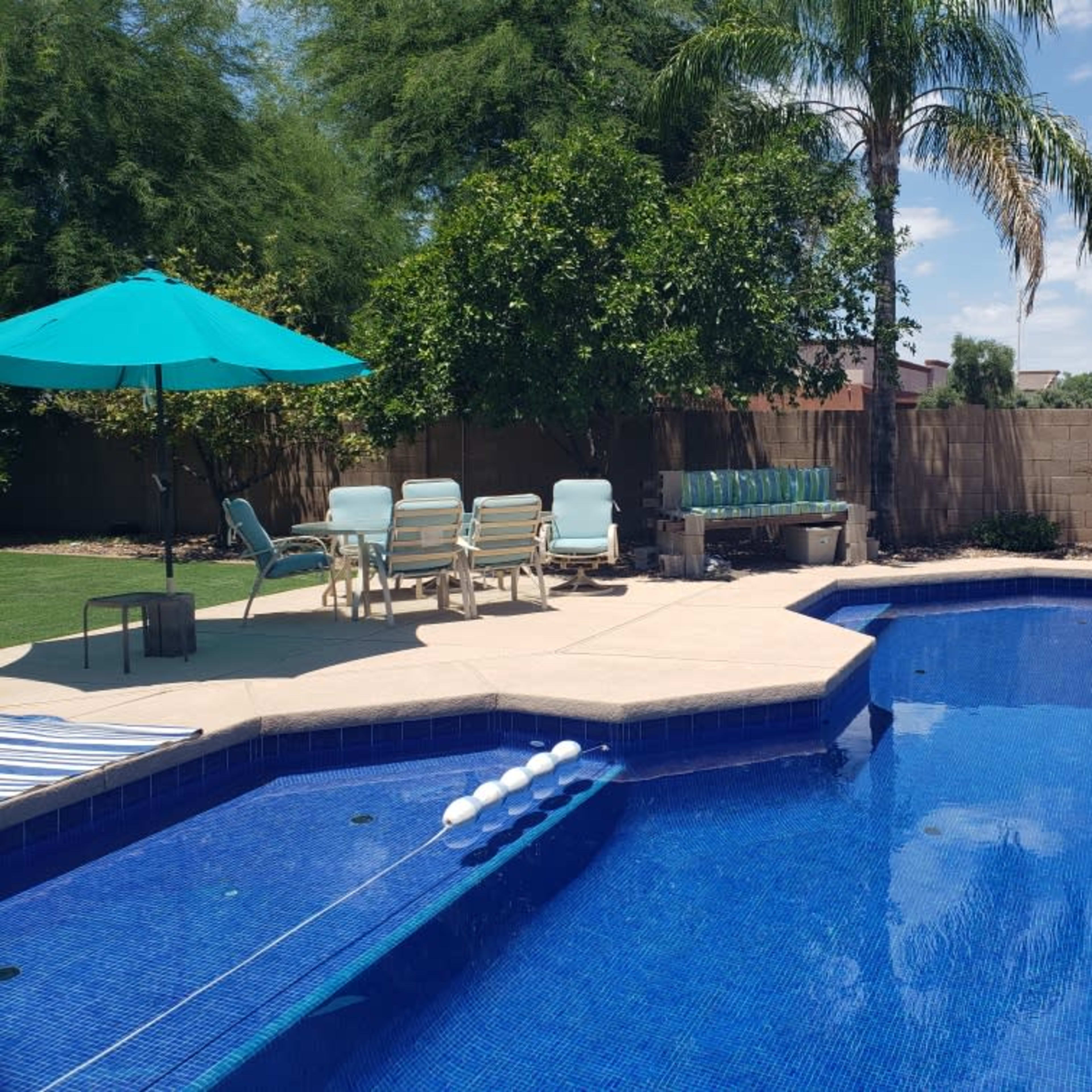 A swimming pool with a basketball hoop, surrounded by lounge chairs and an umbrella in a backyard setting.
