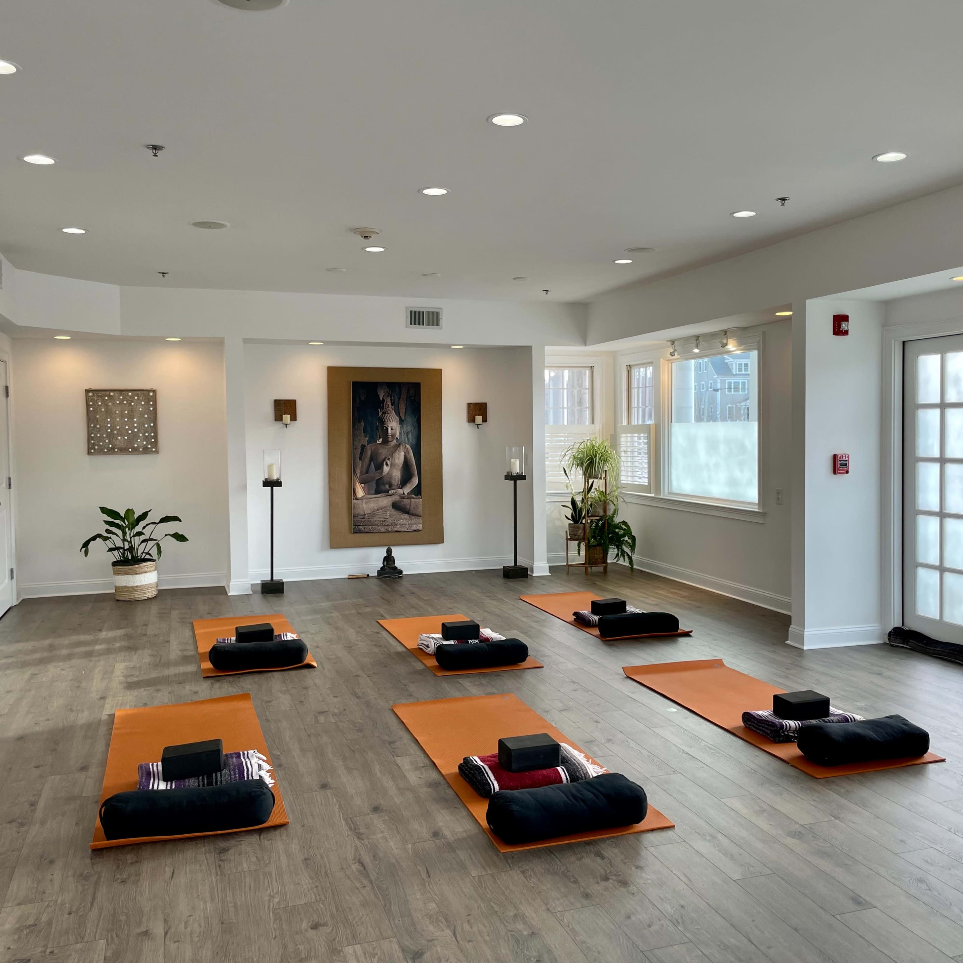 The room features six orange mat setups arranged on a wooden floor, with a large artwork and plants in the background.
