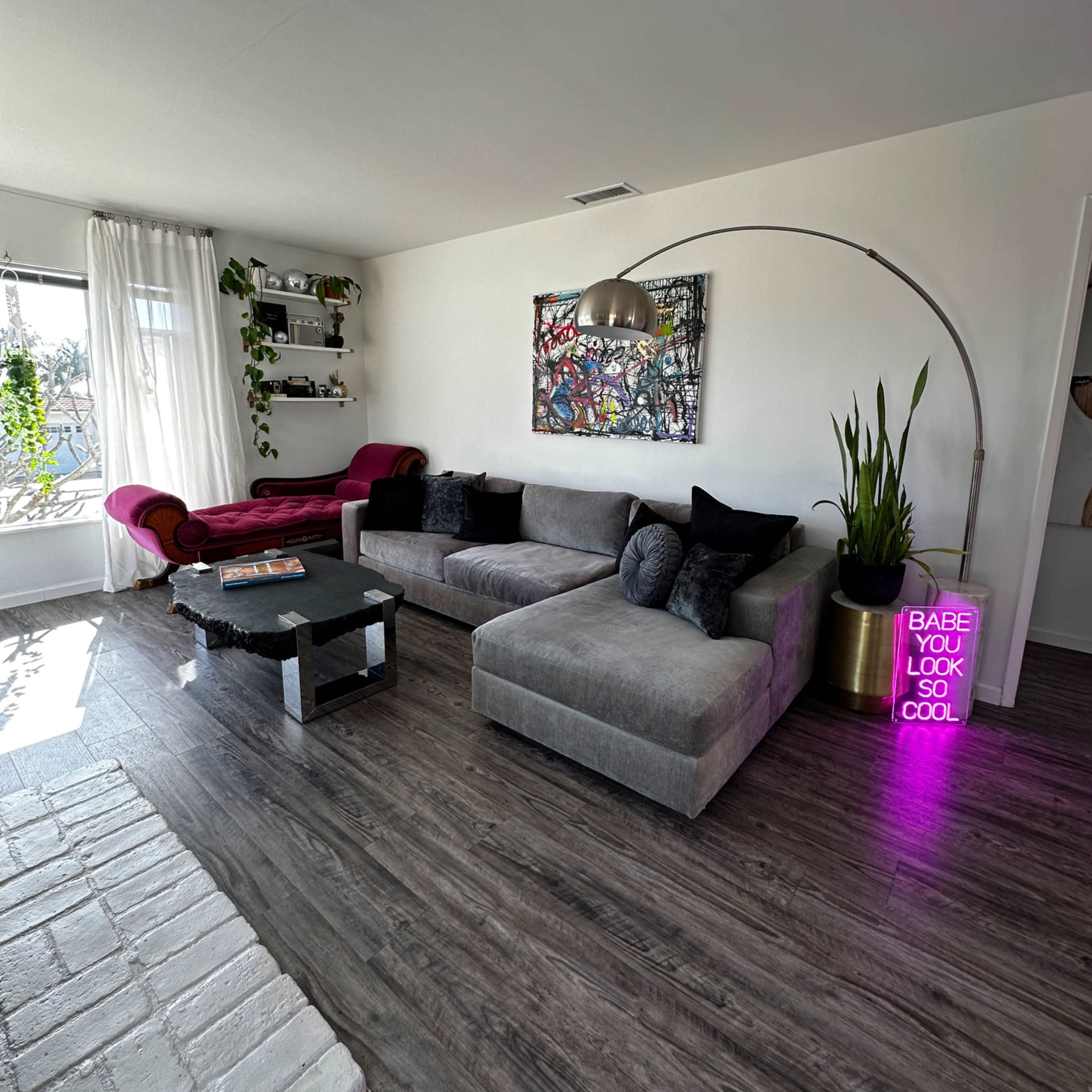 The living room features a large gray sectional sofa, a black coffee table, and a colorful wall art piece, with a neon sign and potted plant in the corner.