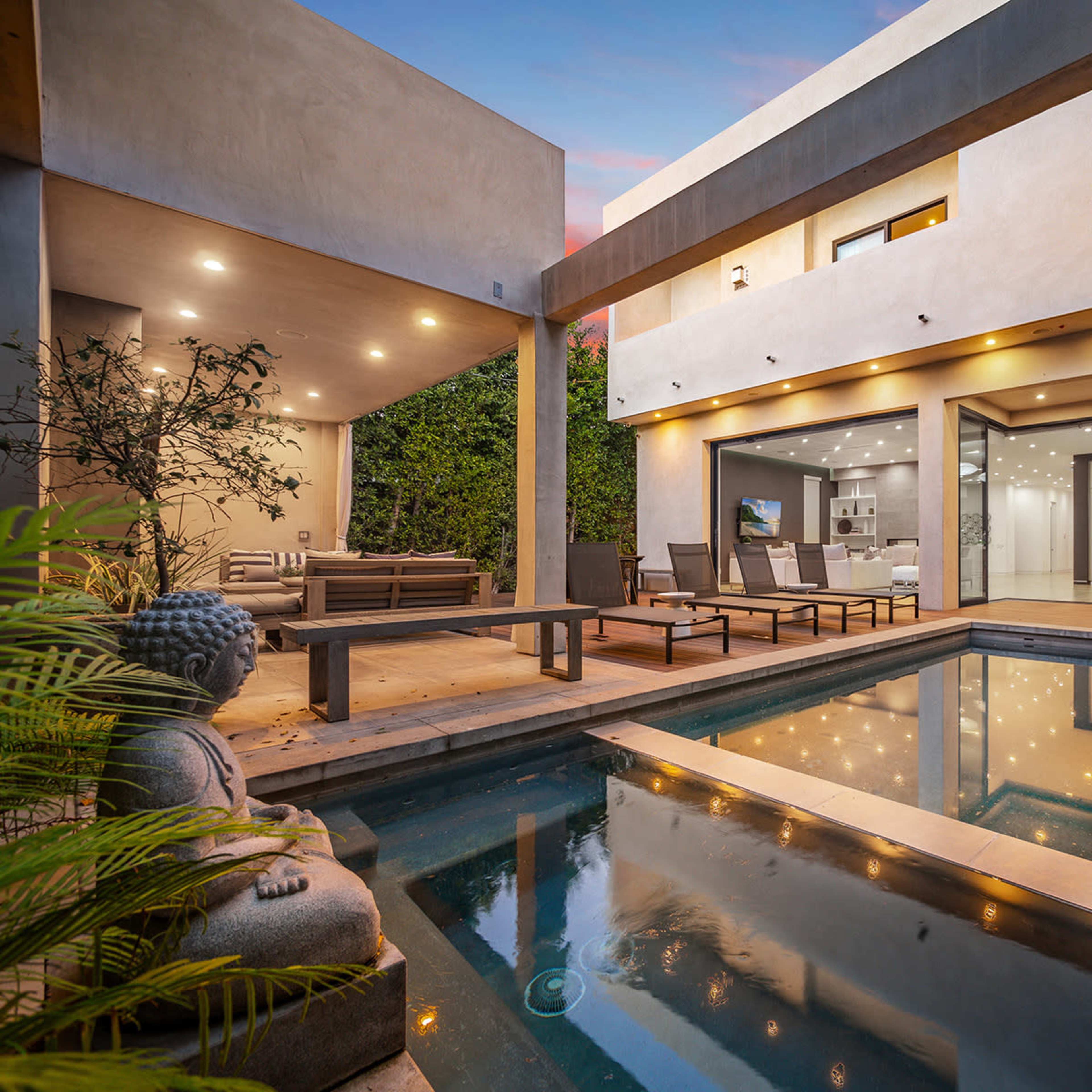 A modern outdoor space featuring a swimming pool, lounge chairs, and a statue, with an adjacent house illuminated by soft lighting.