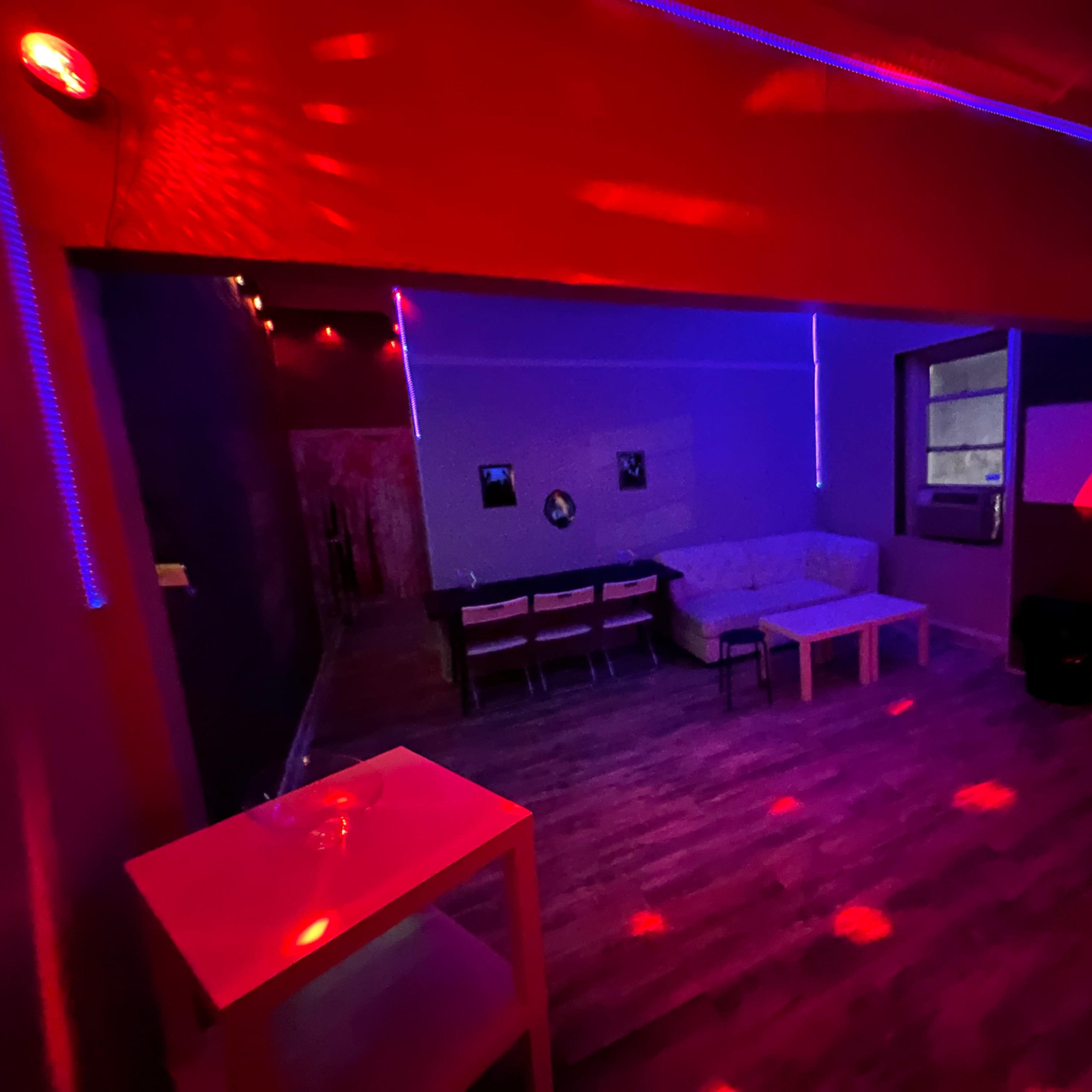 The room features dim lighting with red and blue hues, a white couch, a table, and several chairs arranged in a social setting.
