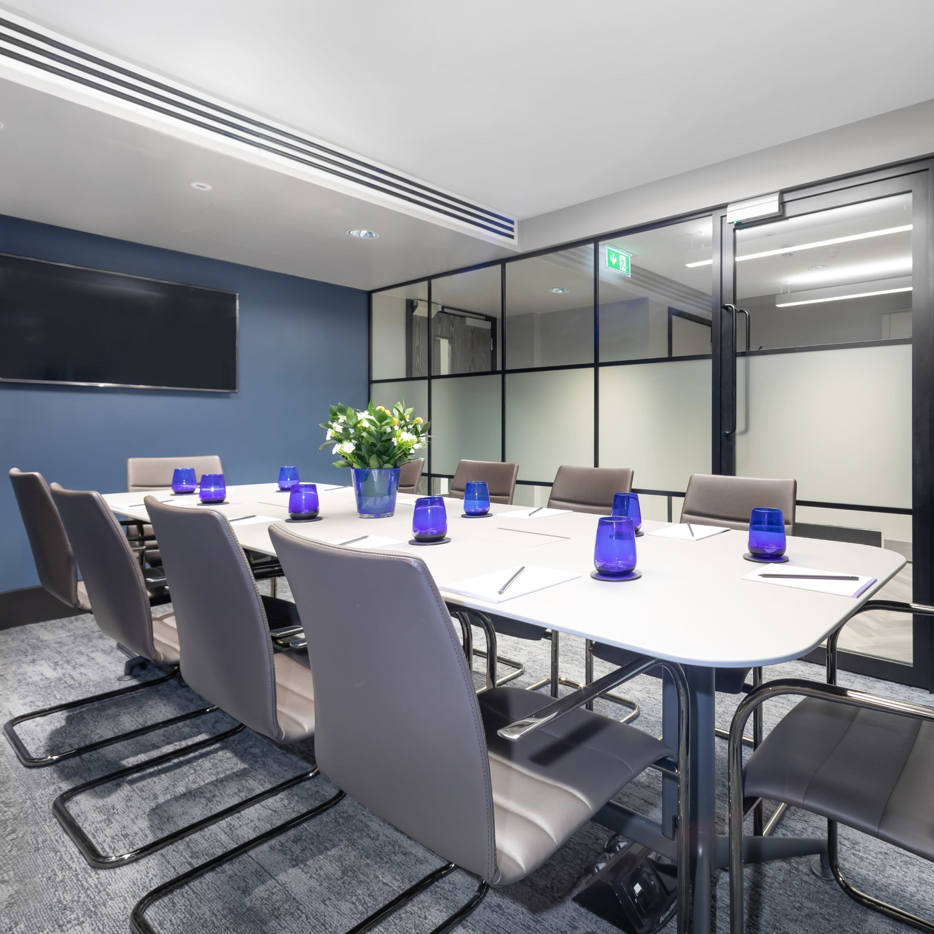 A modern conference room features a large table surrounded by eight ergonomic chairs, with a television mounted on one wall and glass partitions separating it from the hallway.
