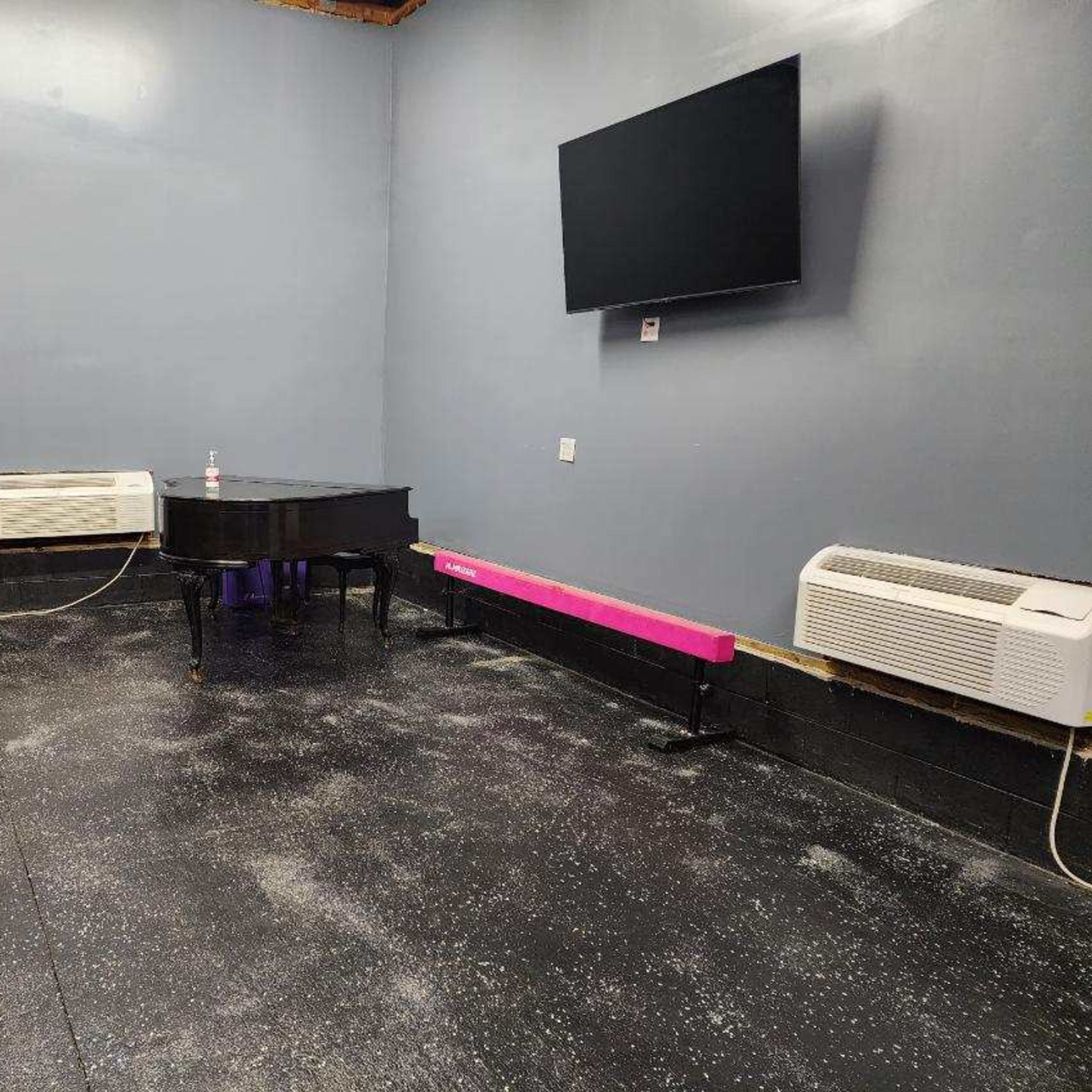 A sparsely furnished room with a grand piano, a black leather couch, a pink bench, and a mounted television on the wall.
