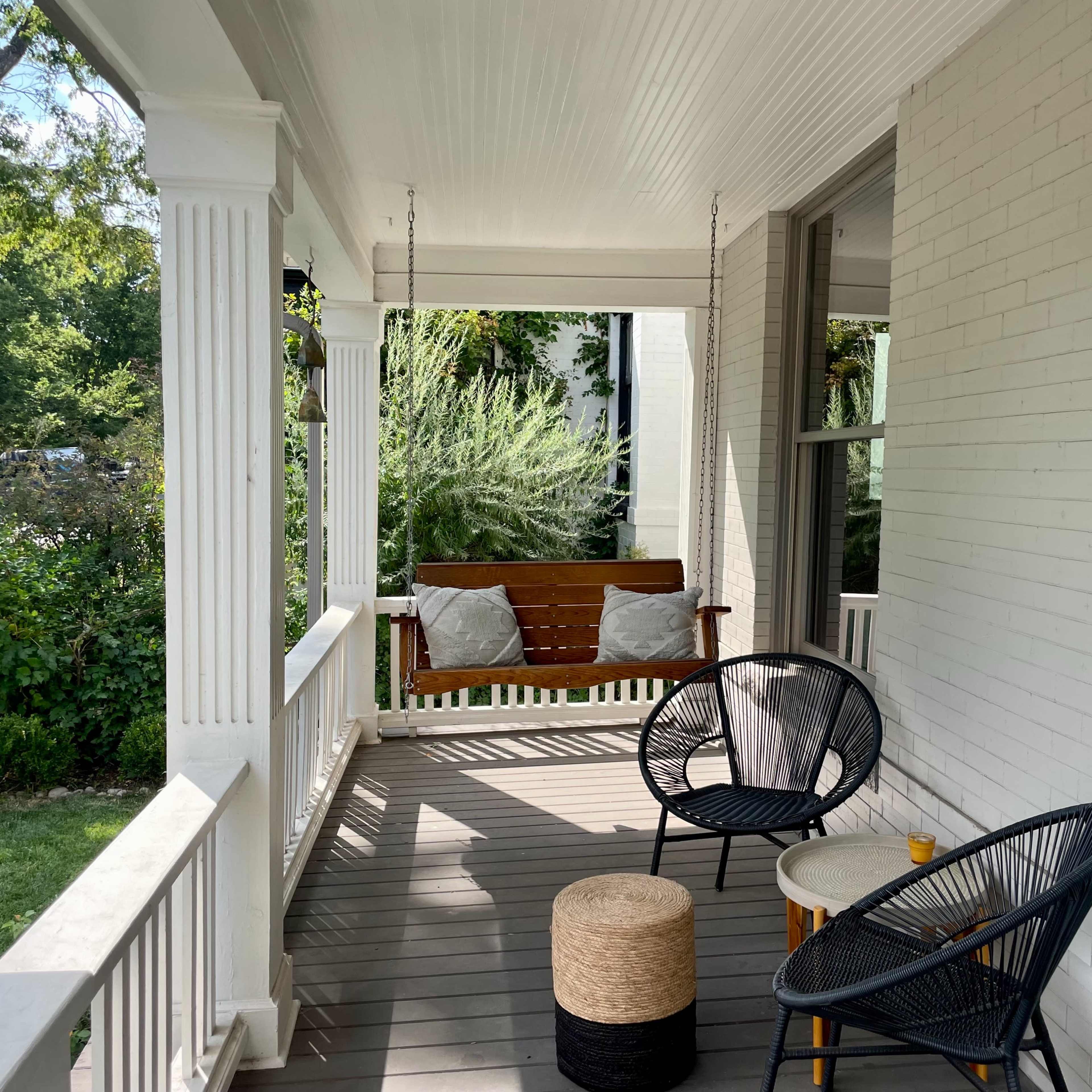 A porch with a swing, two chairs, and a small table, surrounded by greenery.