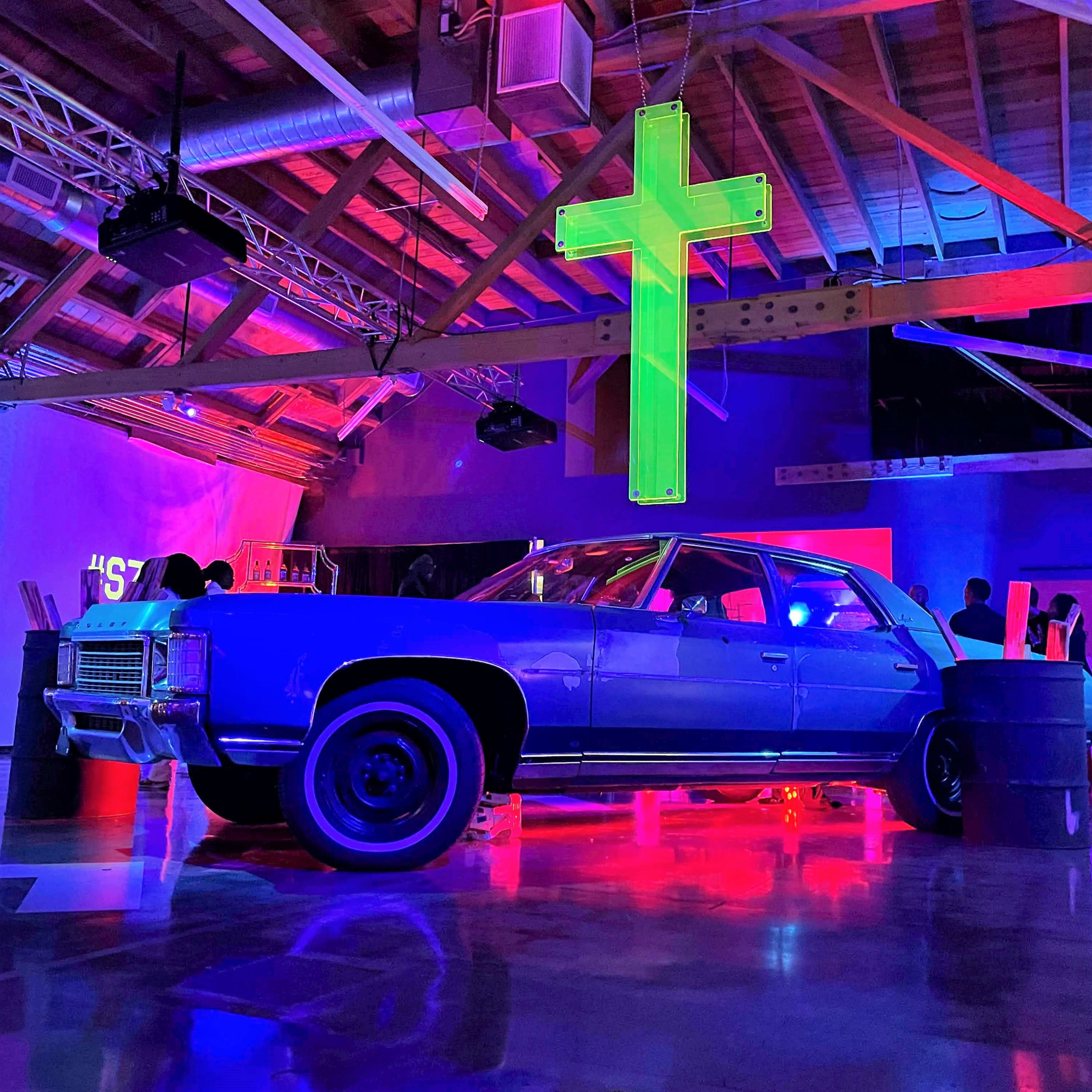 A blue classic car is positioned on a stage under colorful lighting with a large green cross hanging above it in a warehouse-like space.