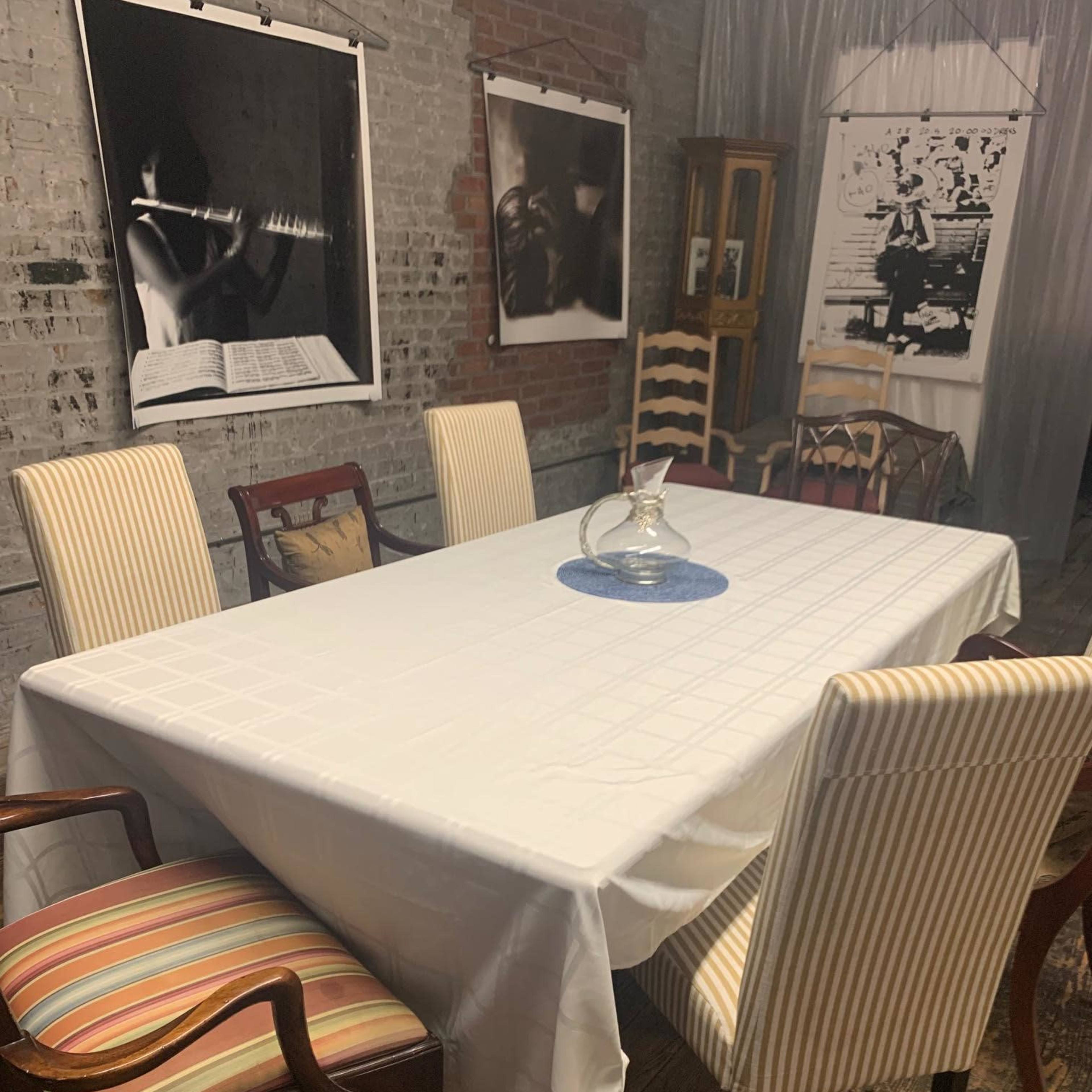 A long, white table is set in a room with brick walls, surrounded by various wooden chairs and framed black-and-white photographs on the walls.