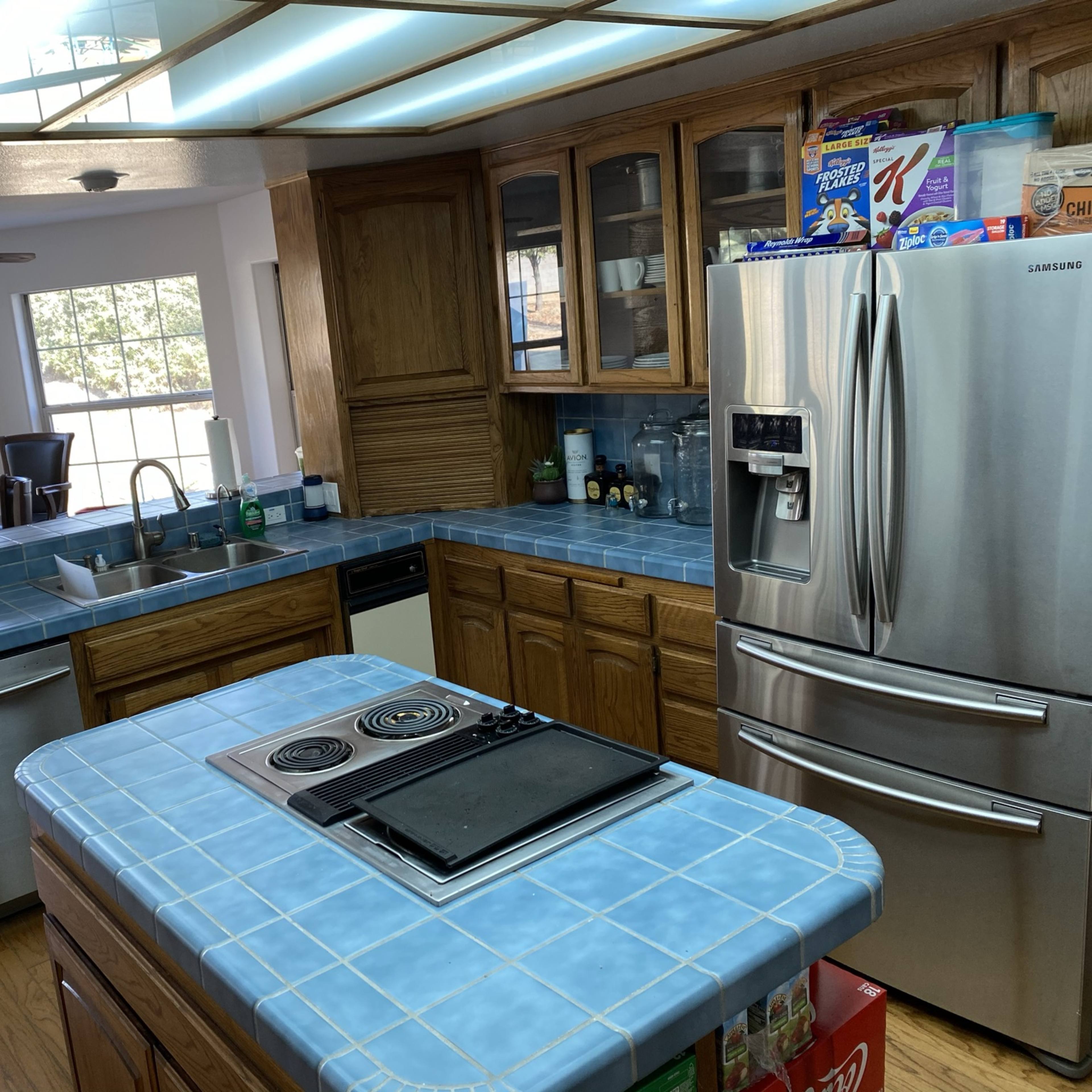 A kitchen with wooden cabinetry, blue tiled countertops, stainless steel appliances, and a central island with a cooktop.