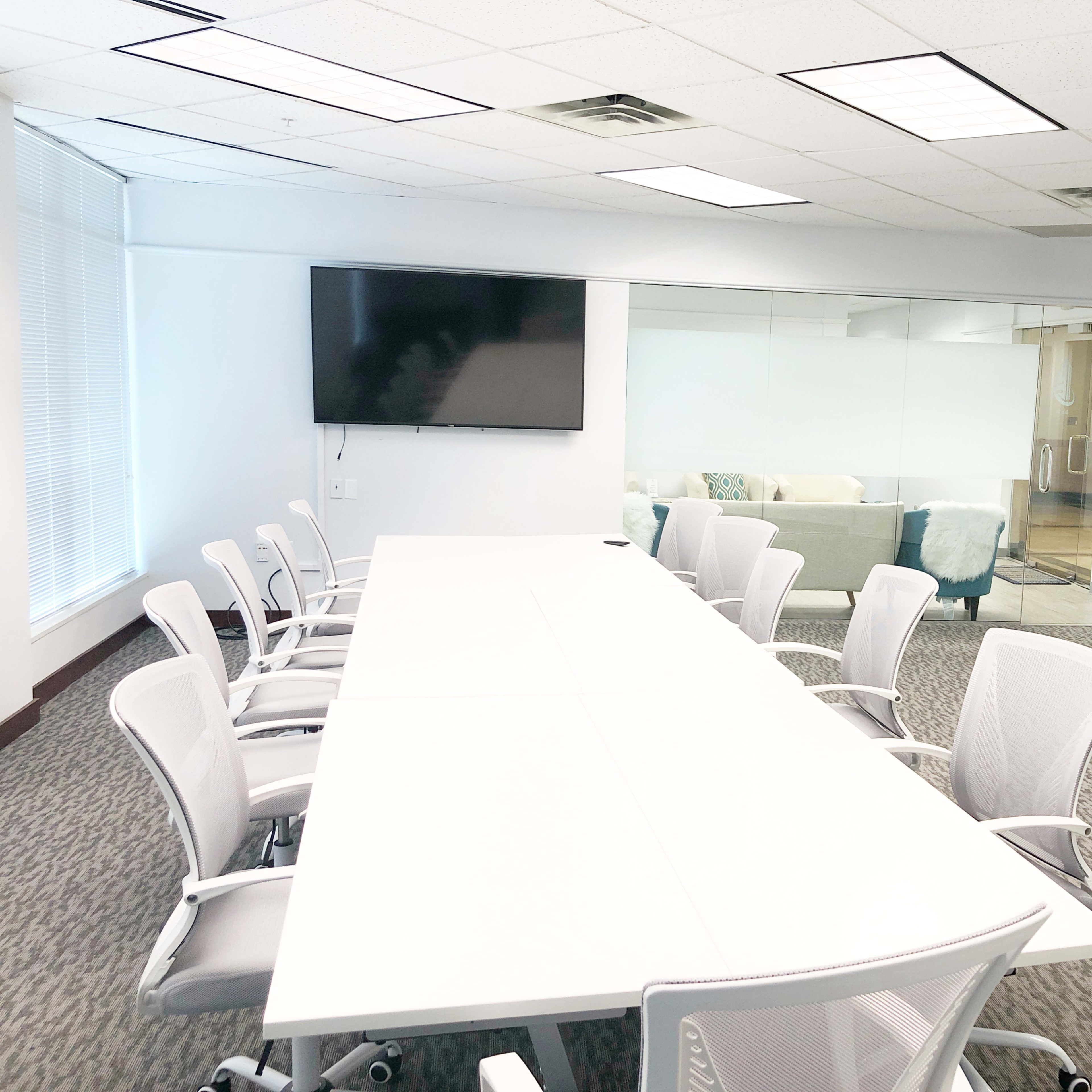 A modern conference room features a long white table surrounded by ergonomic chairs, with a large screen mounted on the wall and a glass partition revealing a separate lounge area.