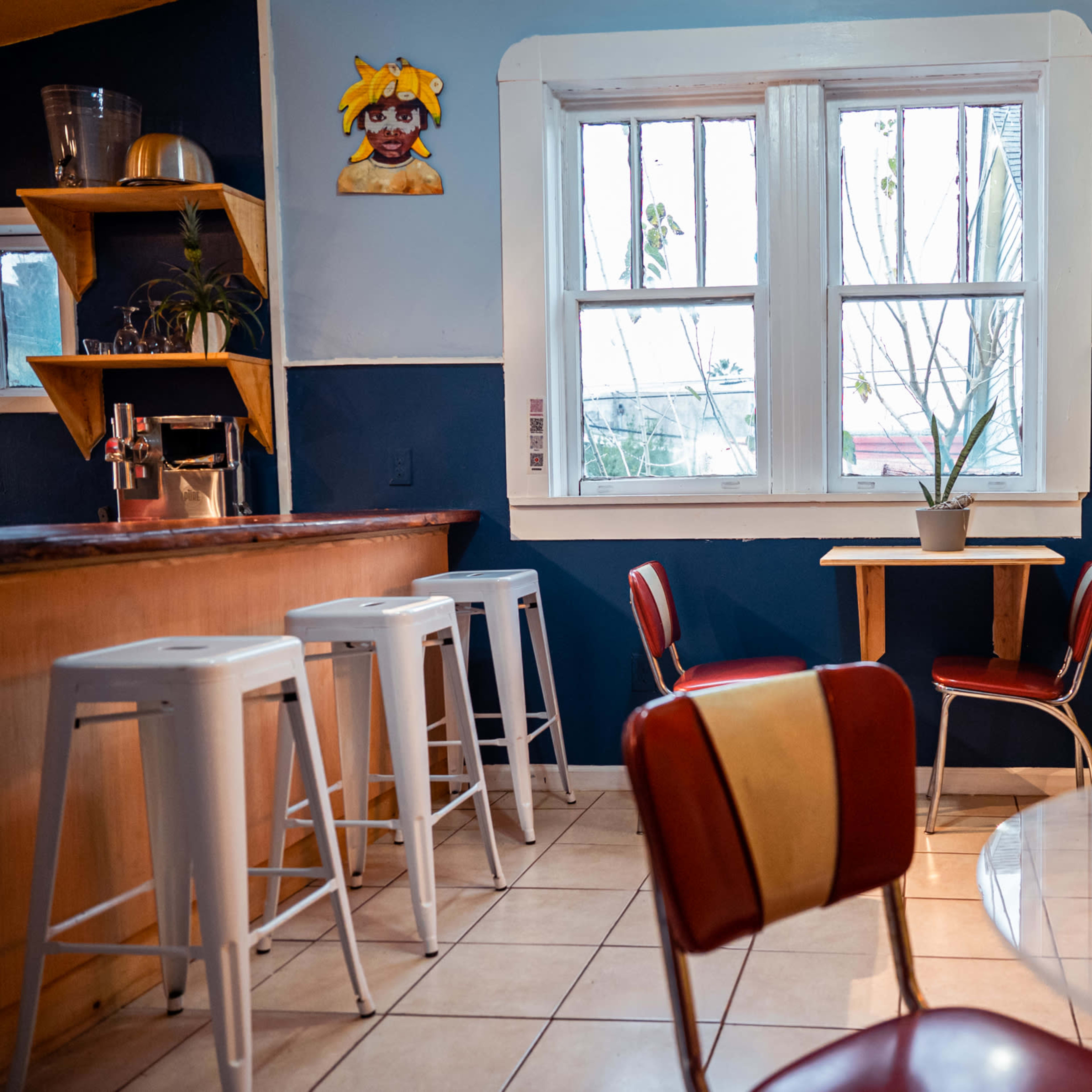A cozy café interior with a bar counter, several stools, and a small table surrounded by red chairs near a window.