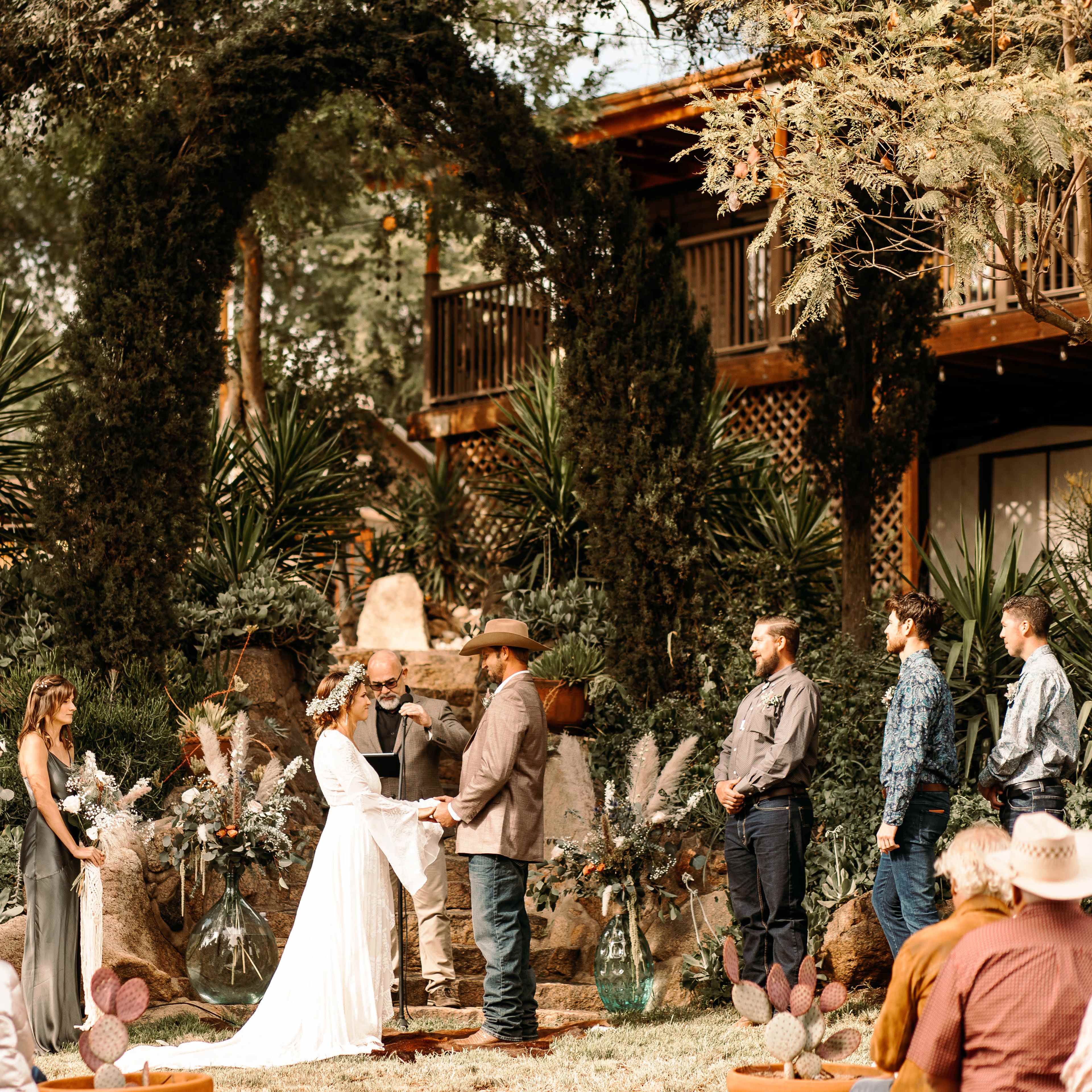 A couple exchanges vows under an arch in a garden setting, surrounded by guests and a wedding party.