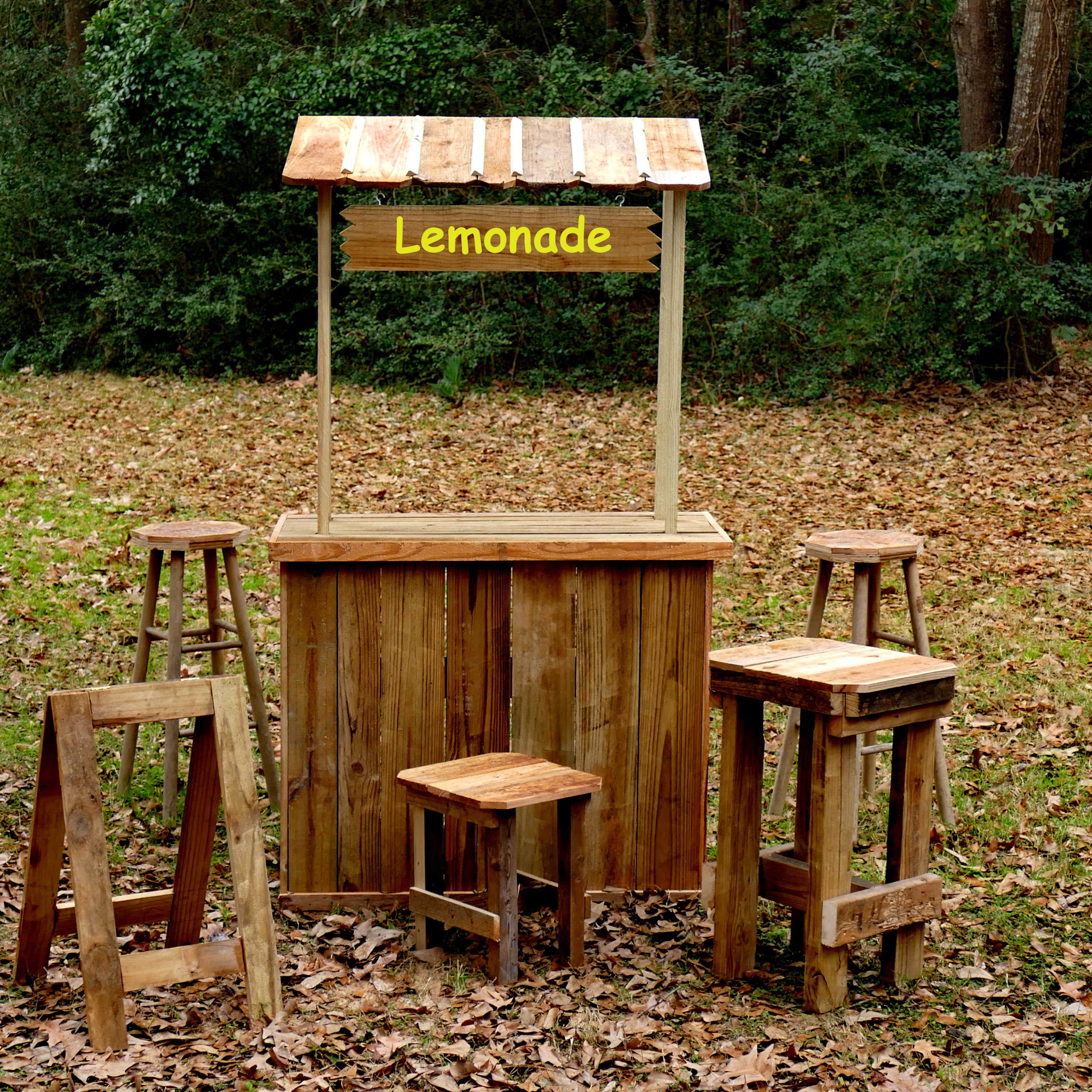A wooden lemonade stand with a slanted roof and five stools is set up in a grassy area covered with fallen leaves.