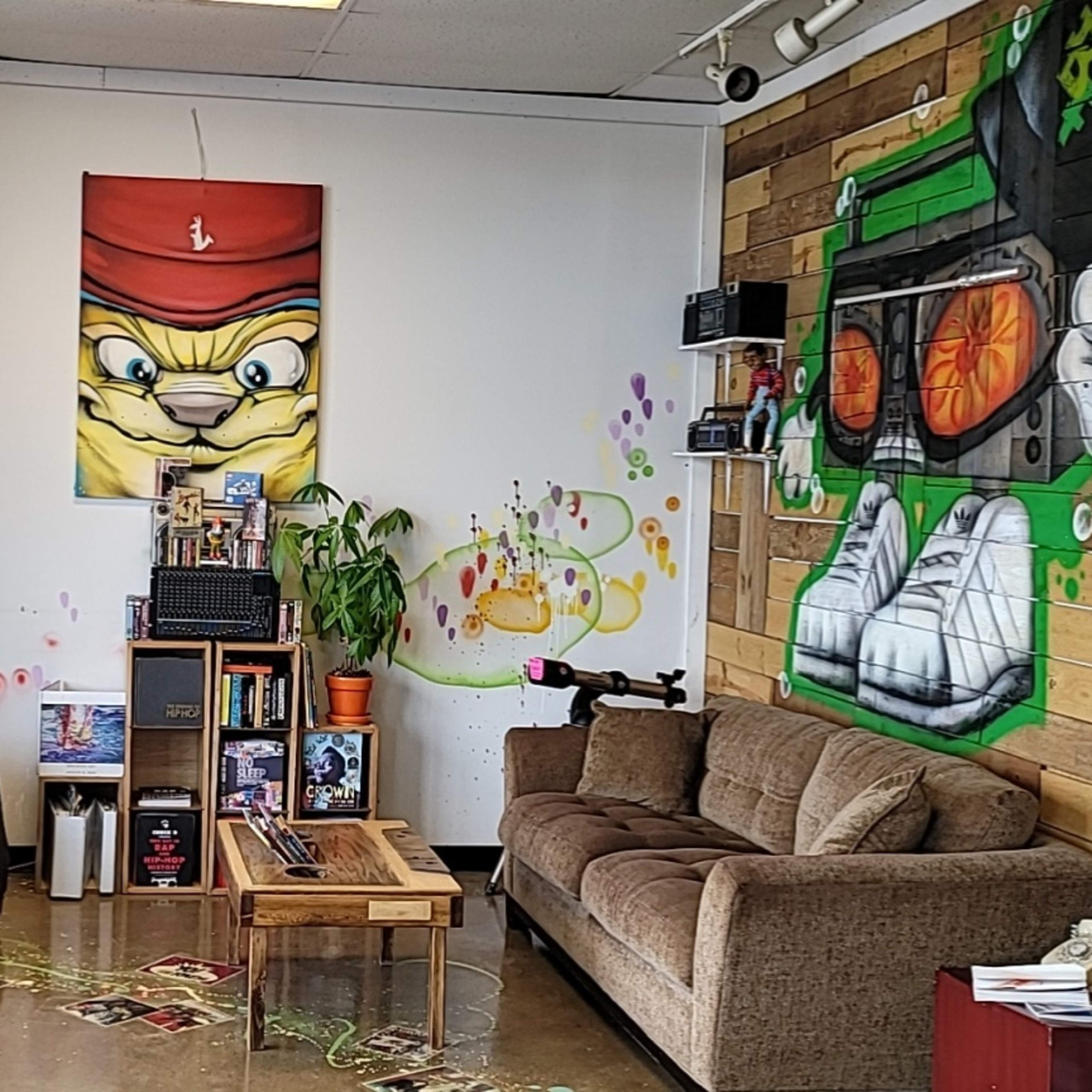 A small lounge area with two gray chairs, a brown couch, a wooden coffee table, and colorful graffiti on the walls, including a large painted character and musical equipment.