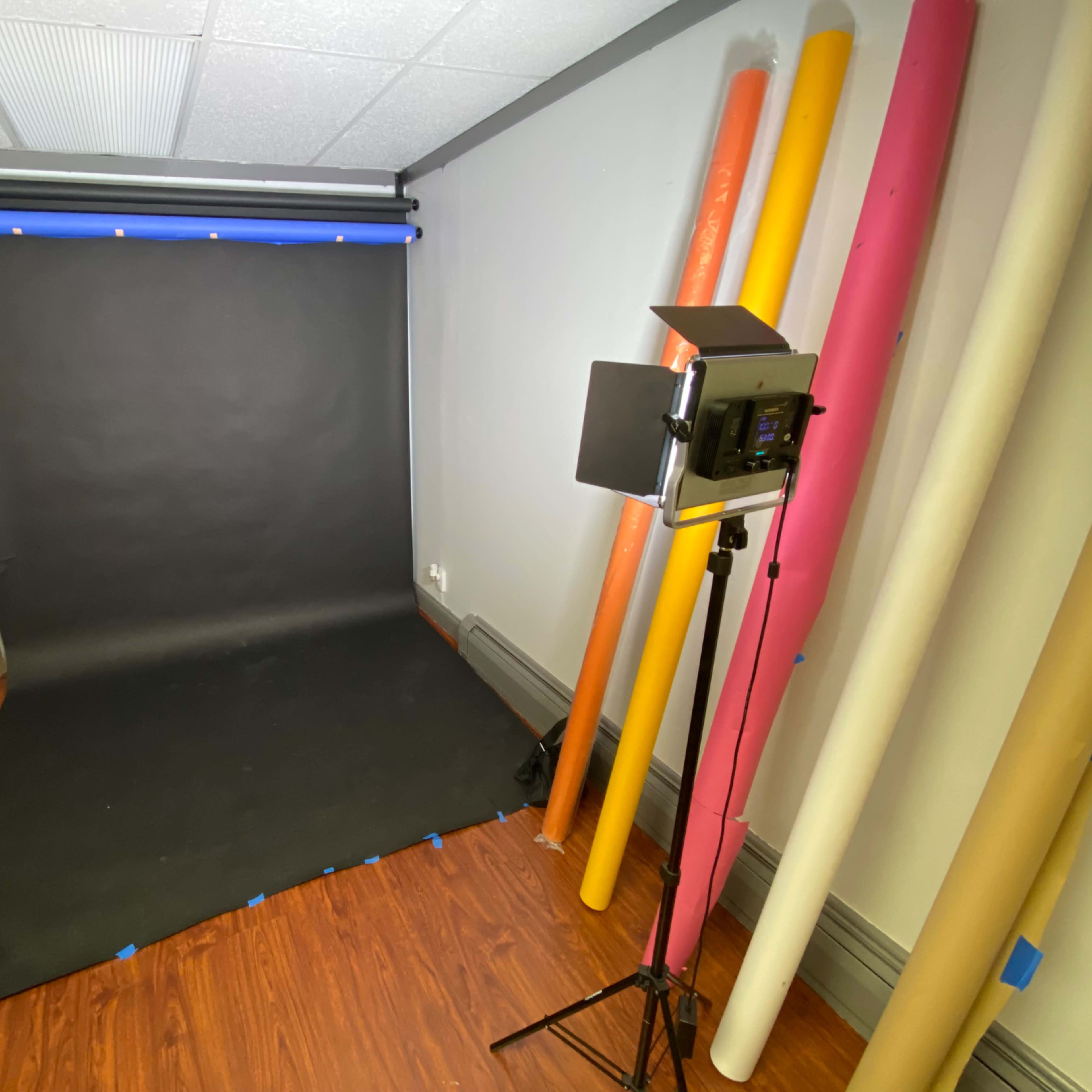 A photography studio setup with a black backdrop, lit by a light source, and various colored rolls of backdrop paper along the wall.