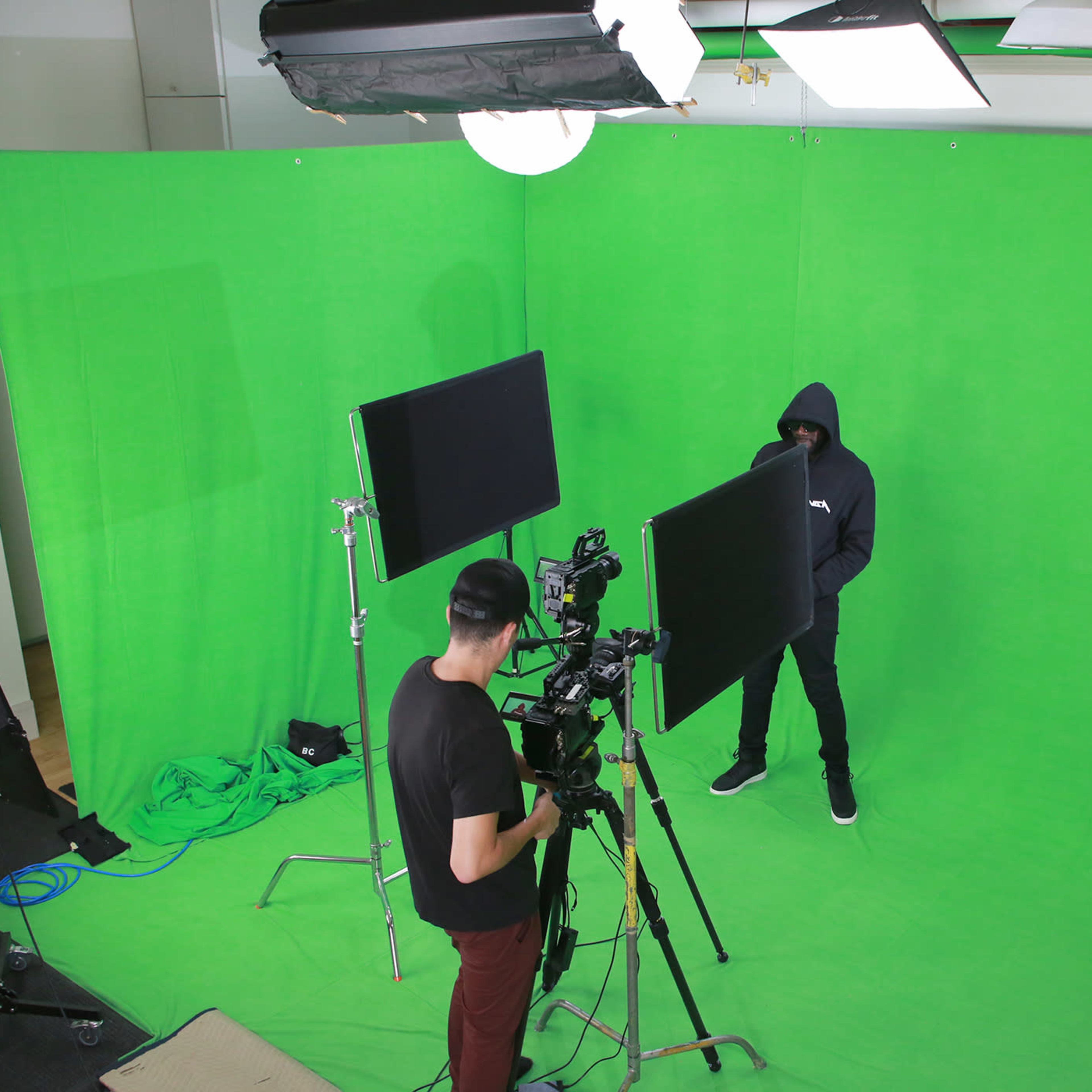 A person in a hoodie stands in front of a green screen while another person operates a camera nearby.