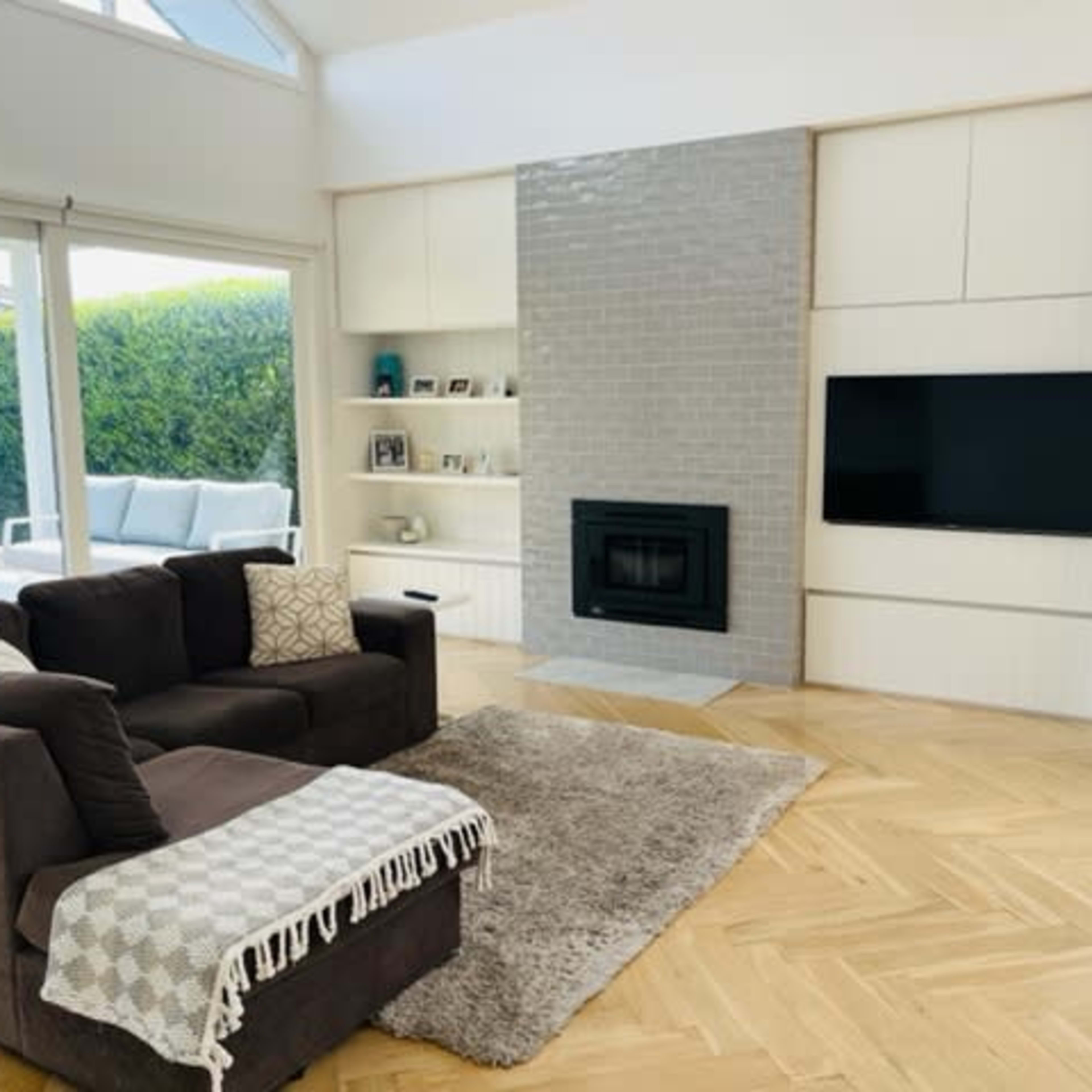 The living room features a gray sofa, a plush rug, a wall-mounted TV, and a fireplace, with large windows revealing an outdoor area.