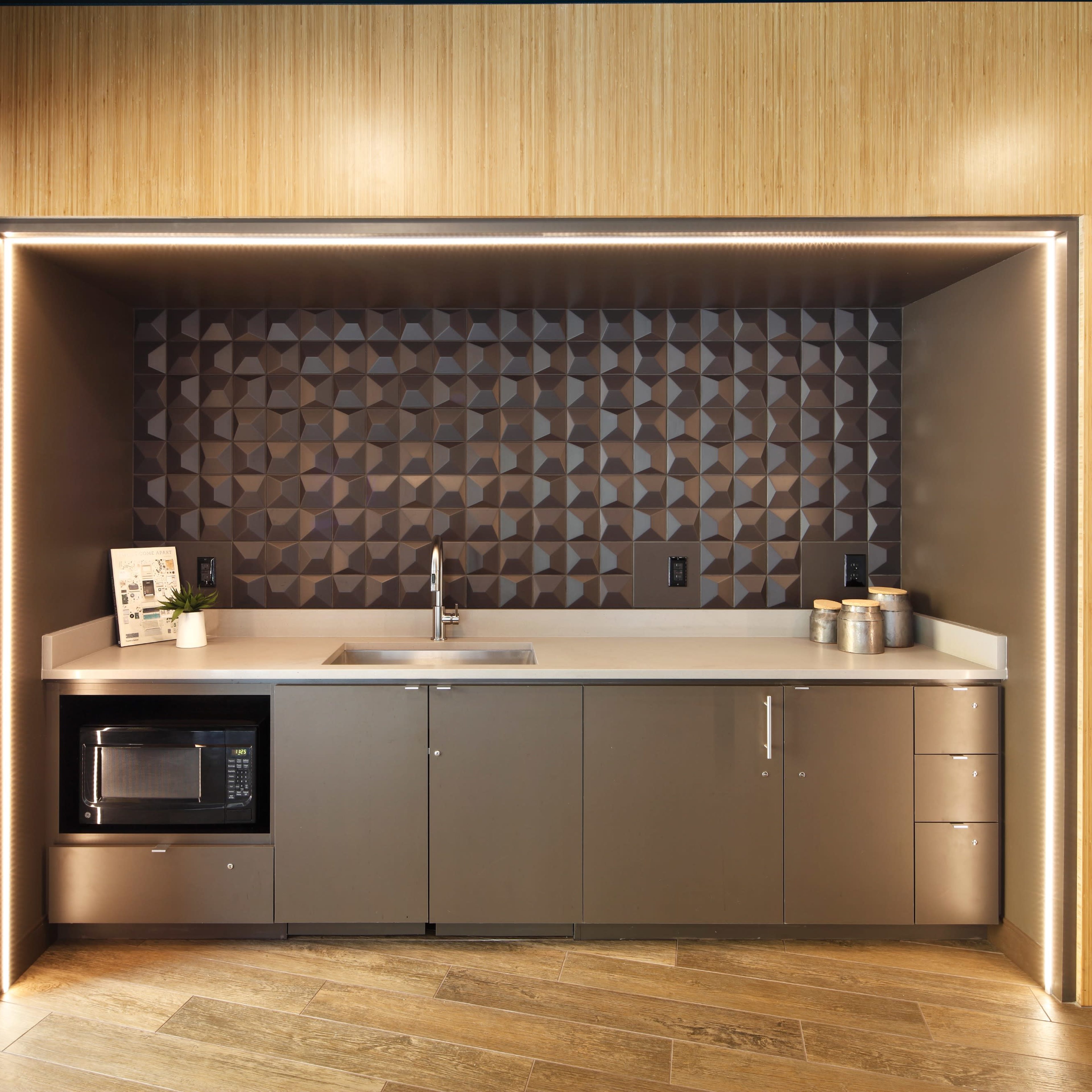 A modern kitchenette features a gray cabinetry with a sink, microwave, and geometric backsplash under soft lighting.