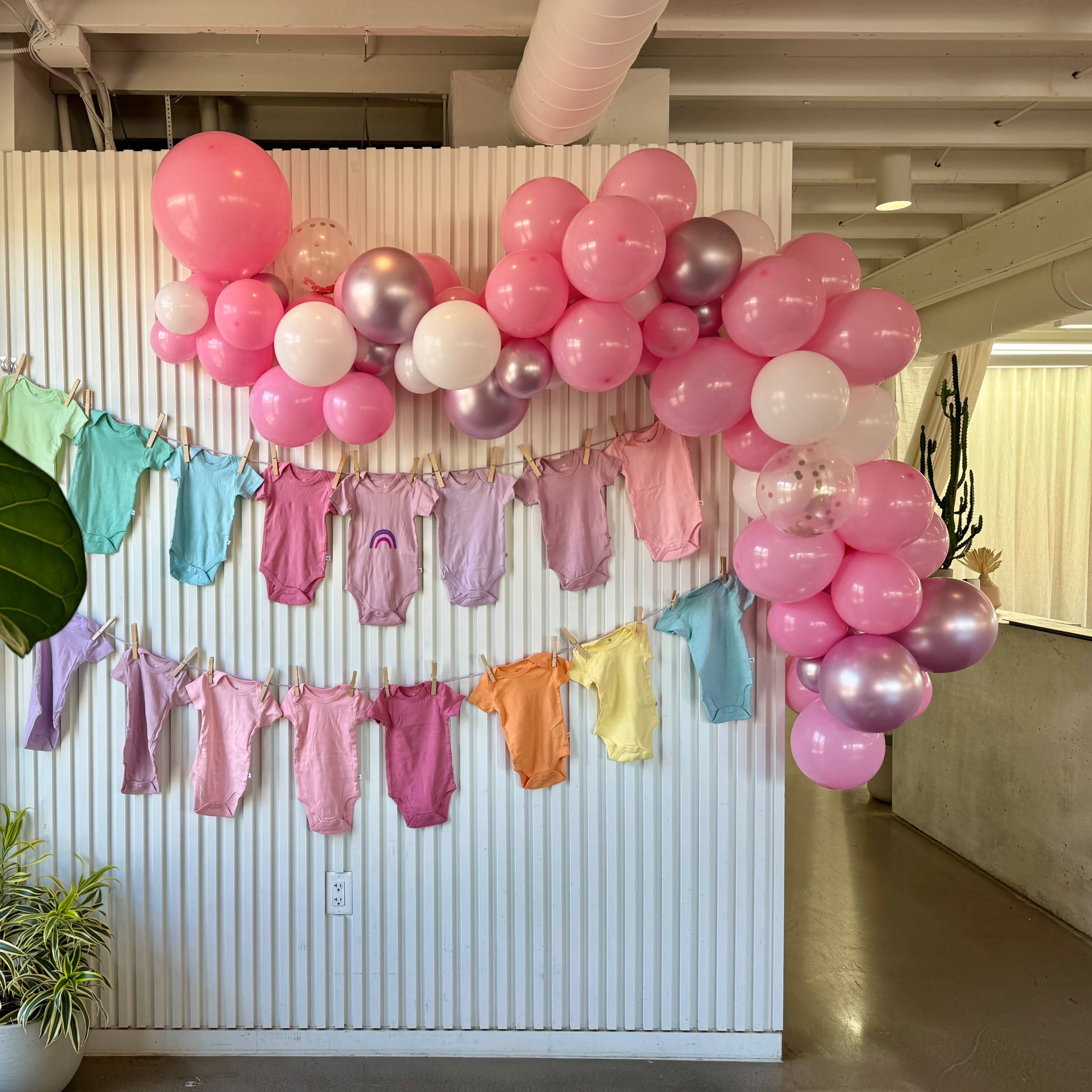 The scene features a wall adorned with pastel-colored baby clothes hanging from a string, complemented by a cluster of pink and white balloons.