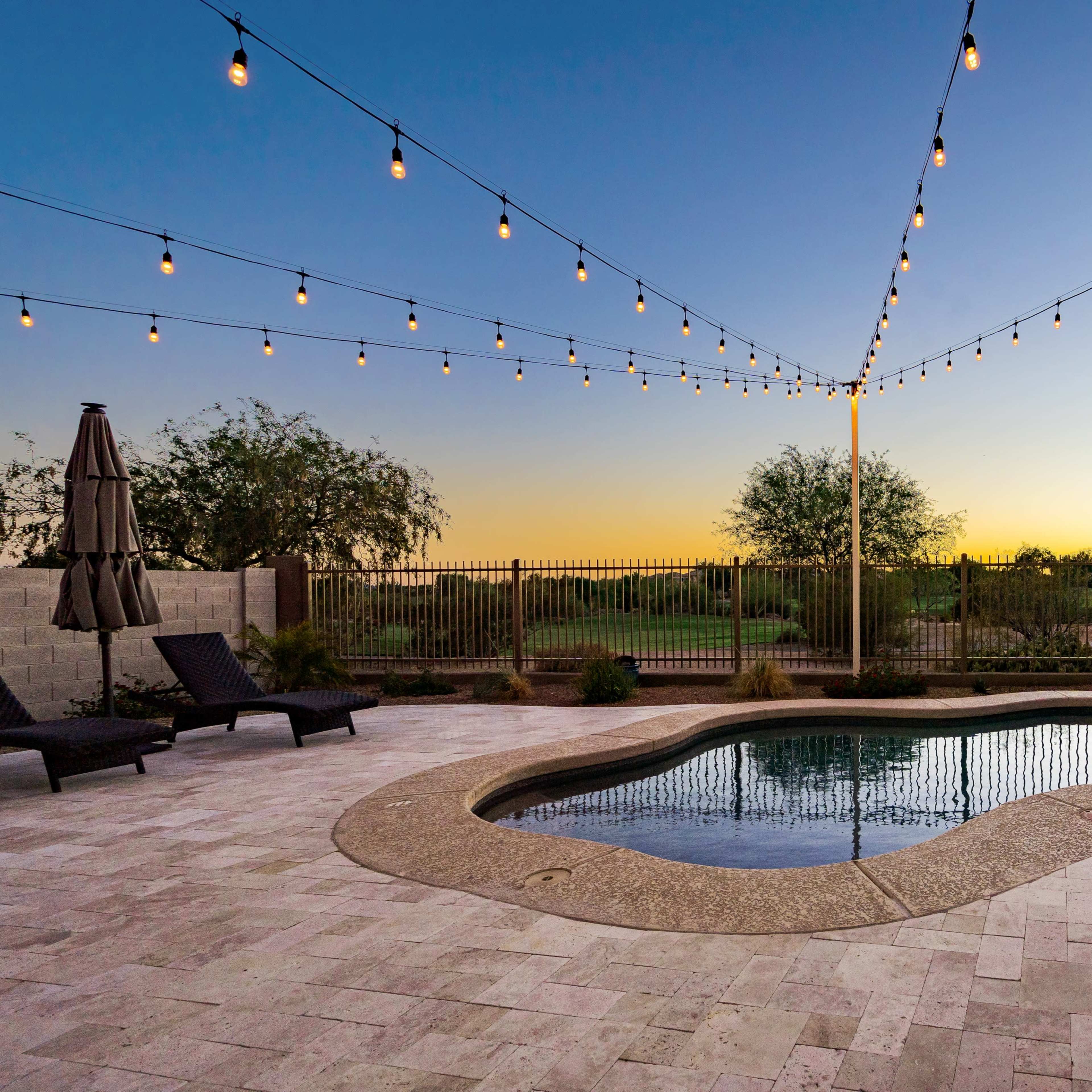 A backyard pool area with a modern design, featuring string lights overhead and a sunset in the background.