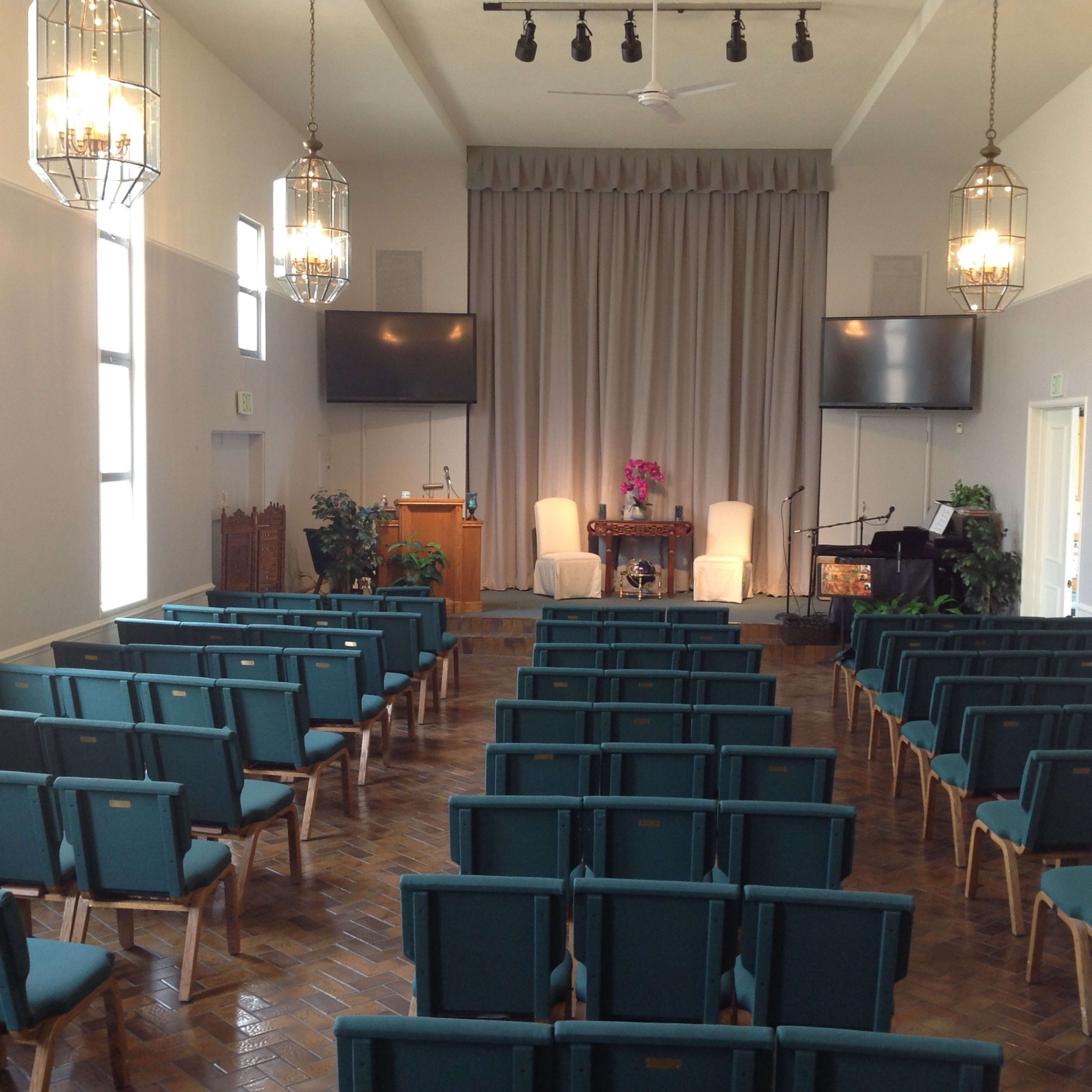 A brightly lit room with rows of teal chairs facing a stage, adorned with a podium, two white chairs, and a piano.
