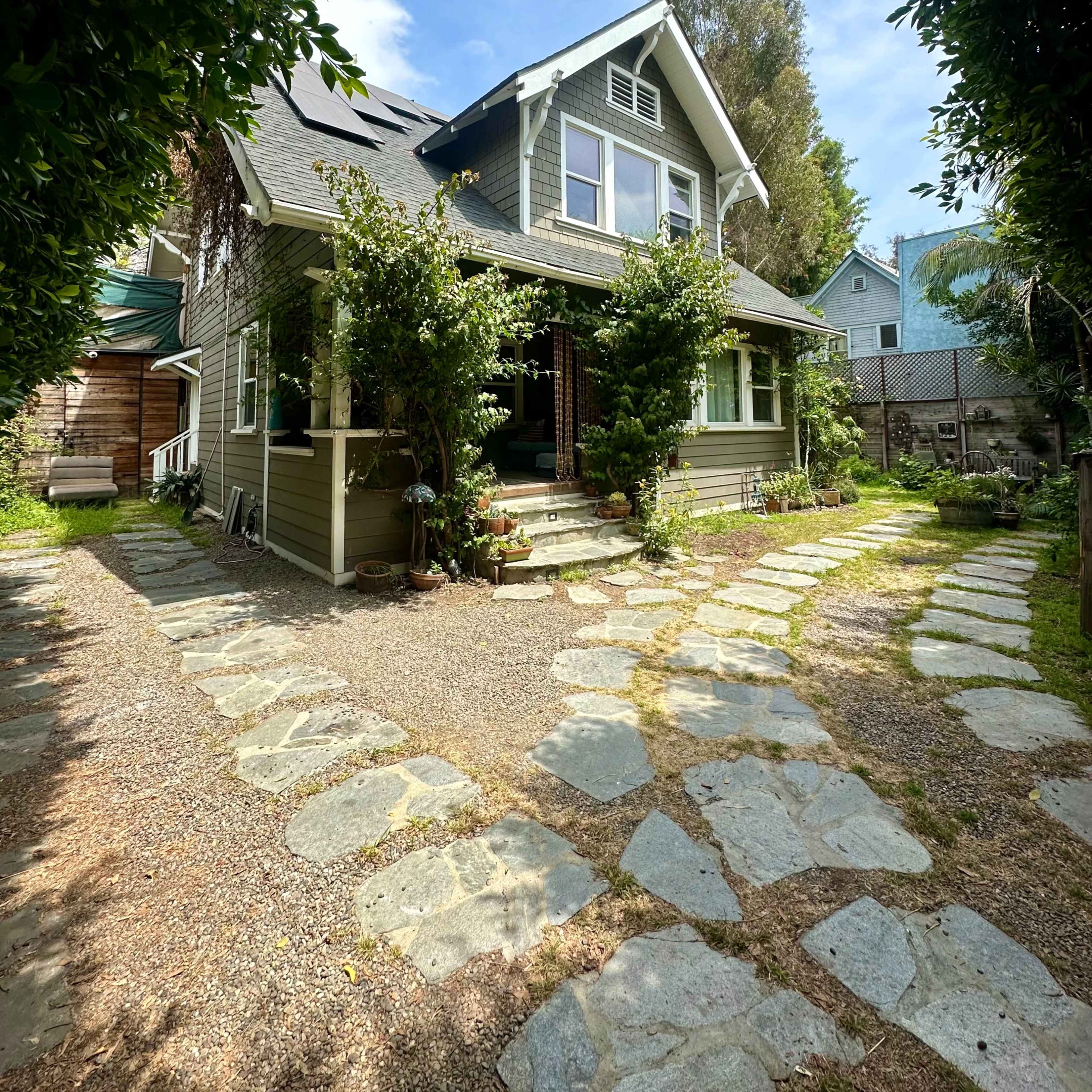 A two-story house with a gabled roof is surrounded by greenery and stone pathways leading to the entrance.