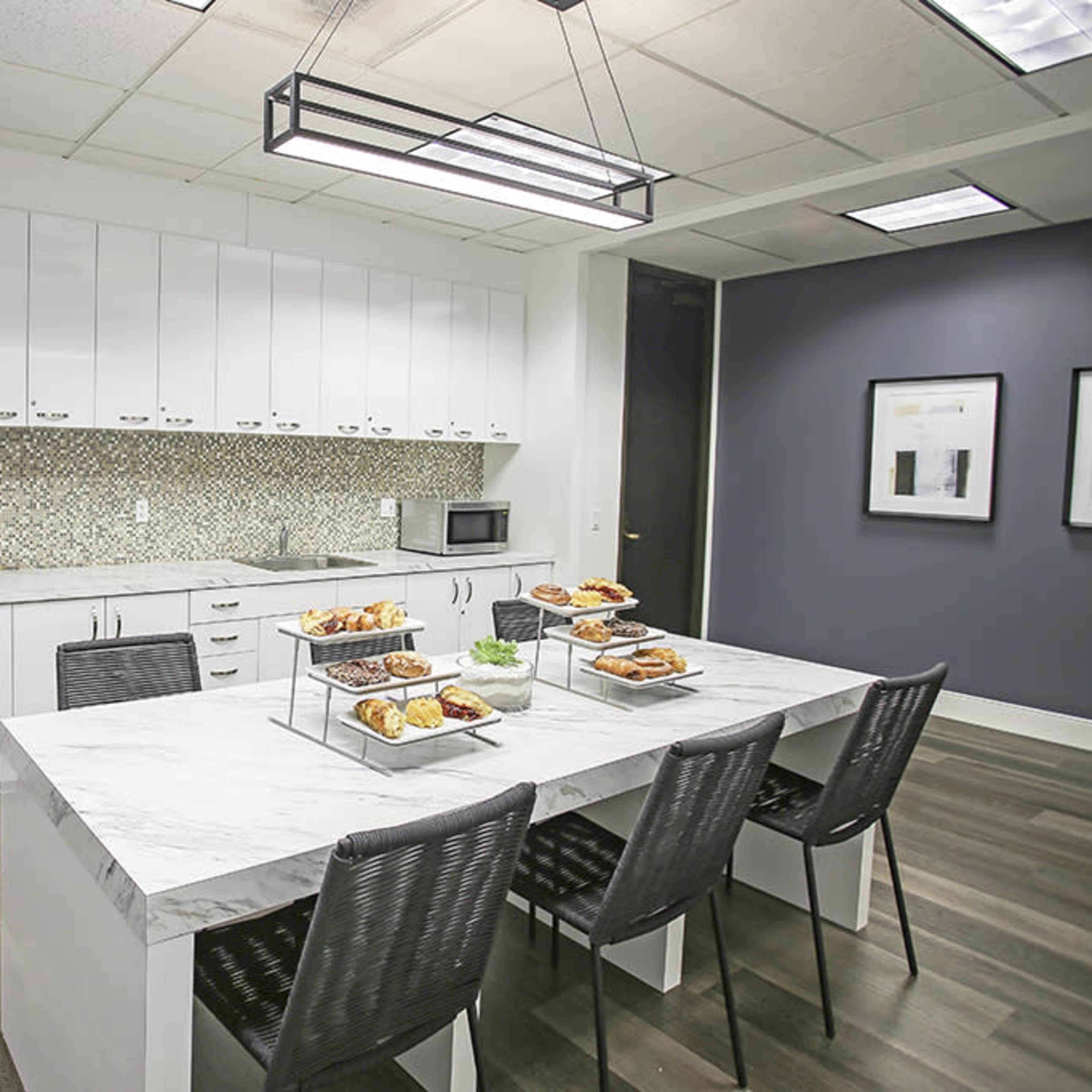 A modern kitchen features a large marble table surrounded by black chairs, with a stainless steel refrigerator, a microwave, and two trays of pastries on the table.