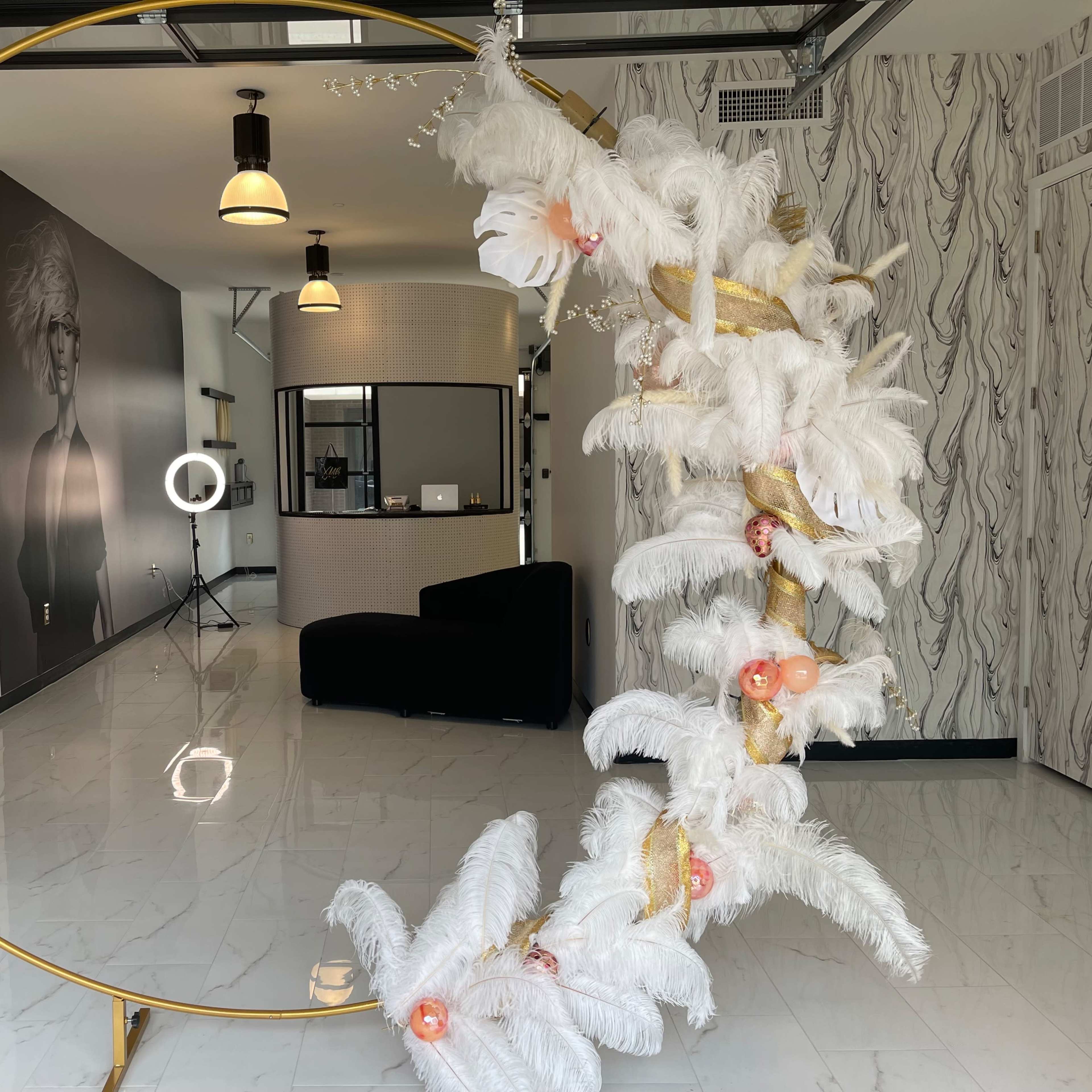 A modern indoor space featuring a decorative gold circular frame adorned with white feathers and floral accents, alongside a reception area with a black sofa and lighting equipment.