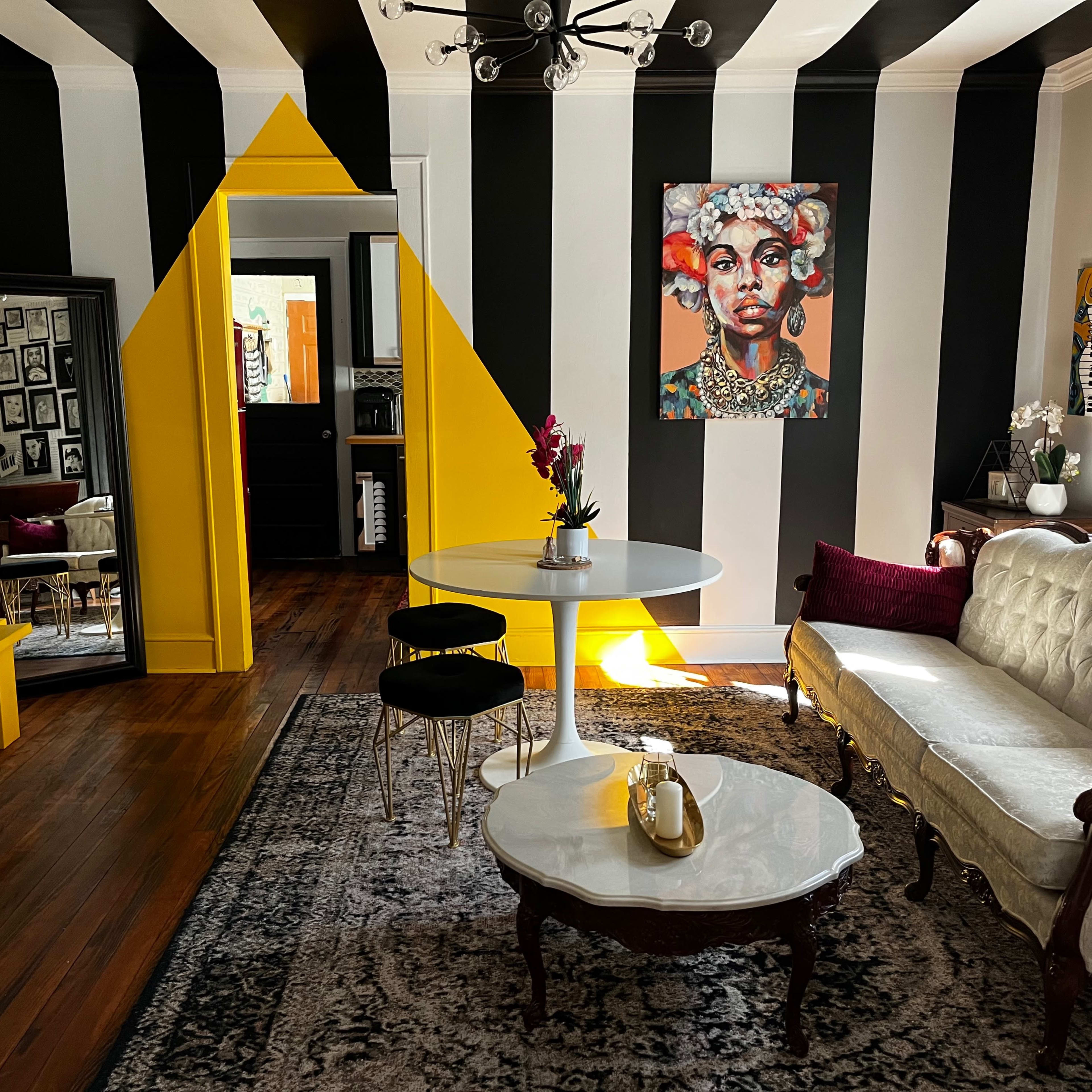 The room features black and white striped walls, a yellow geometric accent wall, a round white dining table, and a vintage sofa, with an artistic portrait displayed above a coffee table.