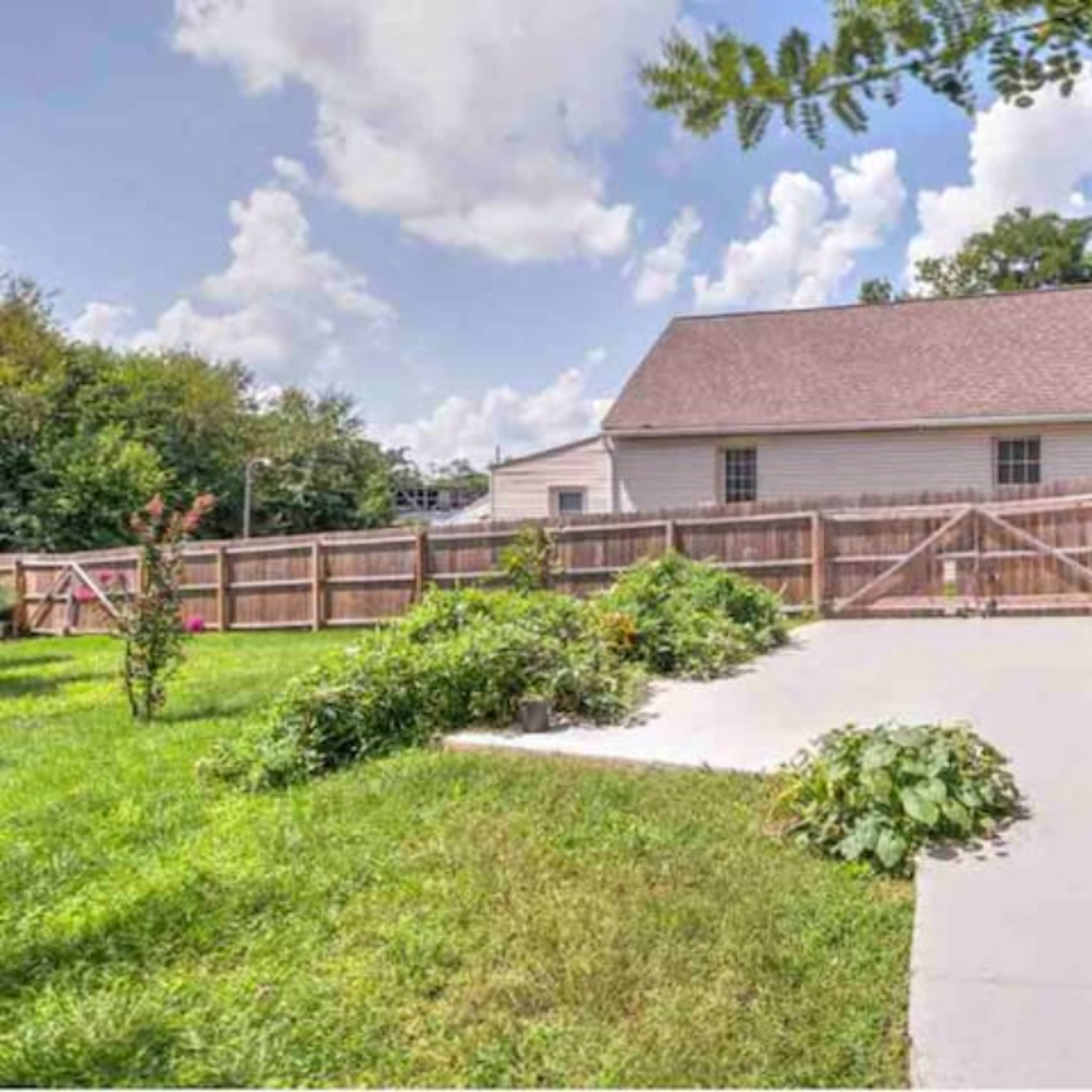 A residential backyard features a concrete pathway leading to a wooden gate, surrounded by grassy areas and shrubs.