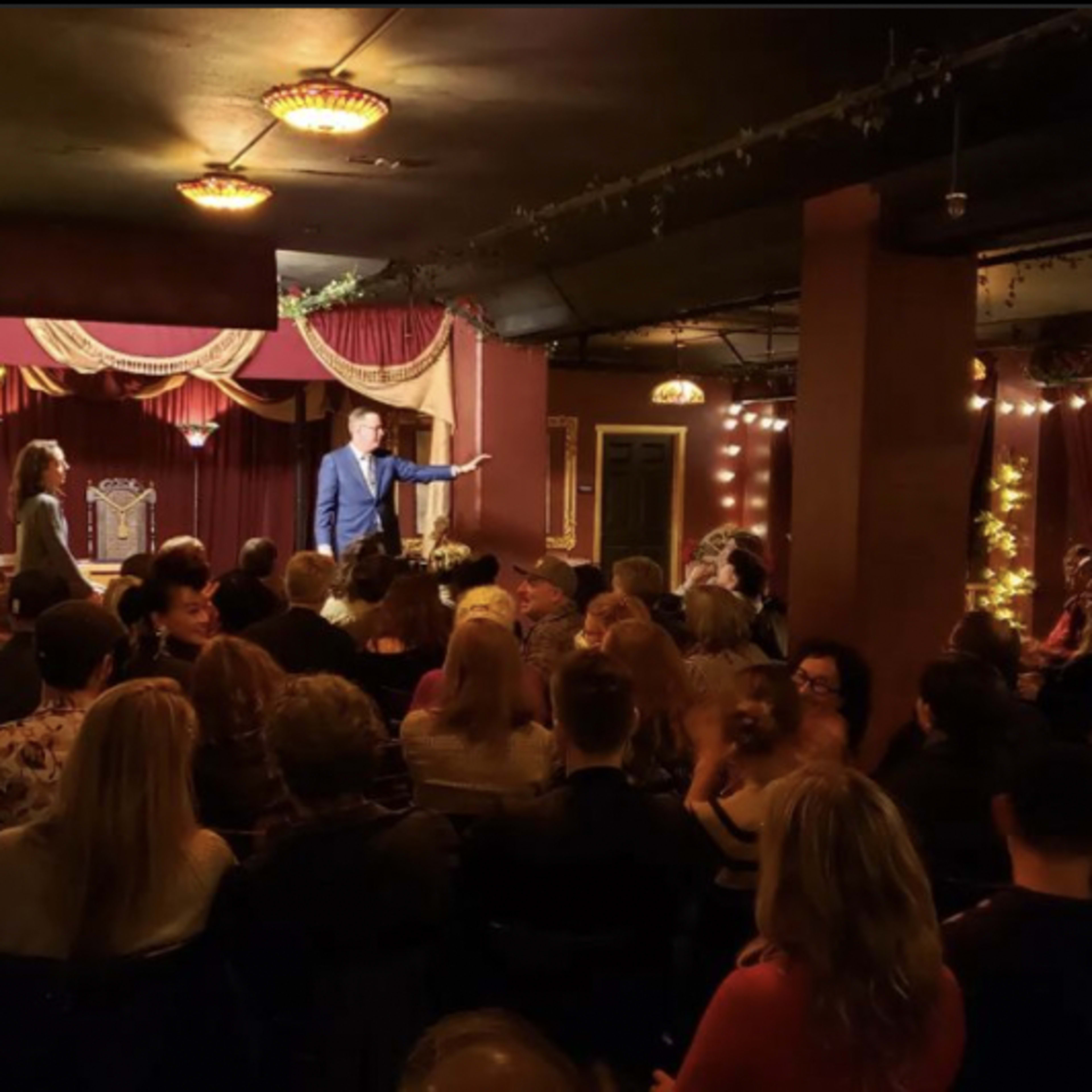 A crowded room with a stage featuring a performer speaking to an audience amidst dim lighting and decorative drapery.