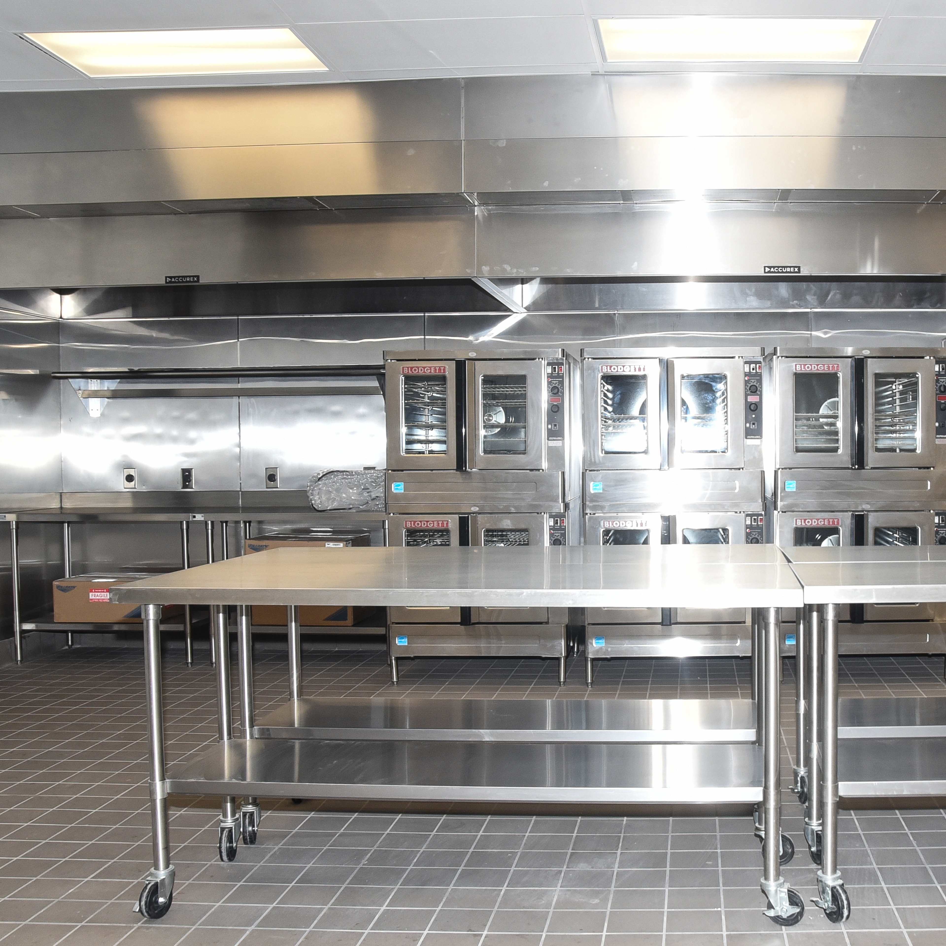 A professional kitchen with stainless steel appliances and countertops, featuring several ovens and open shelving.