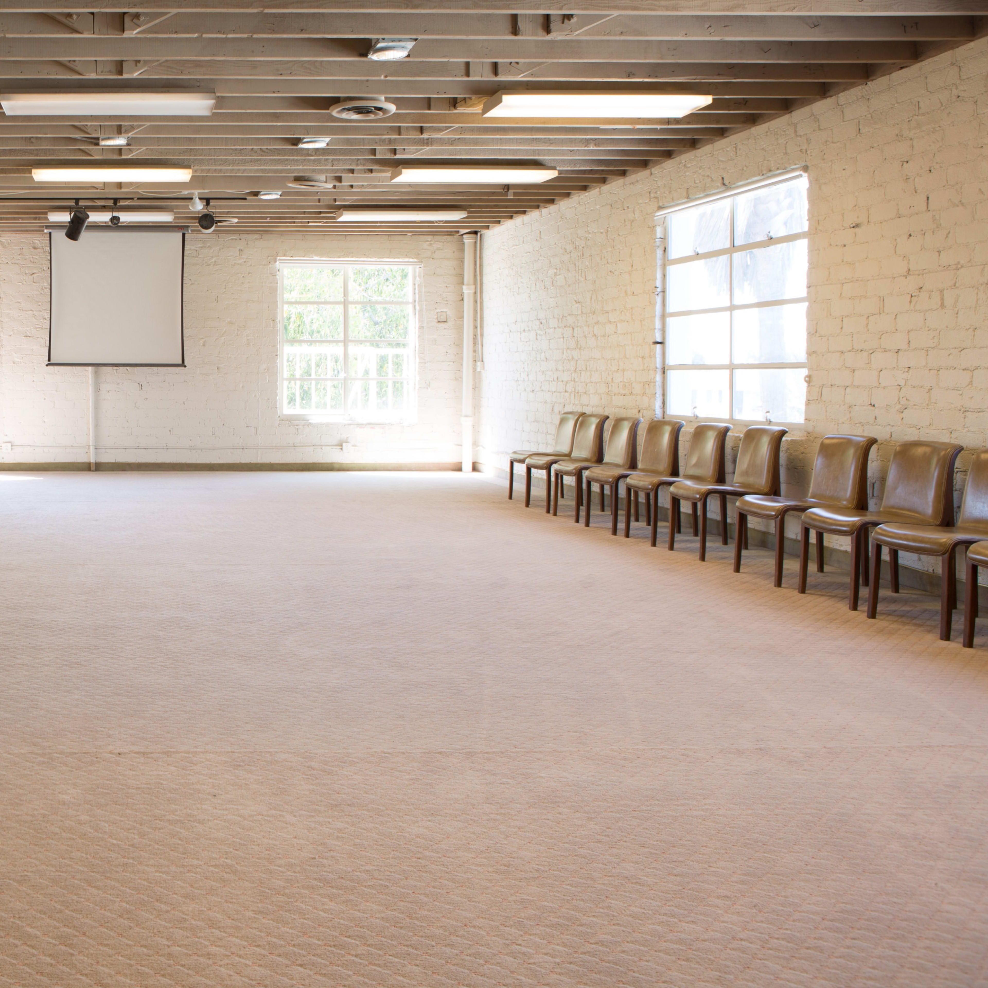 A spacious, empty room with wooden beams on the ceiling, a piano on one side, and rows of chairs along the walls.