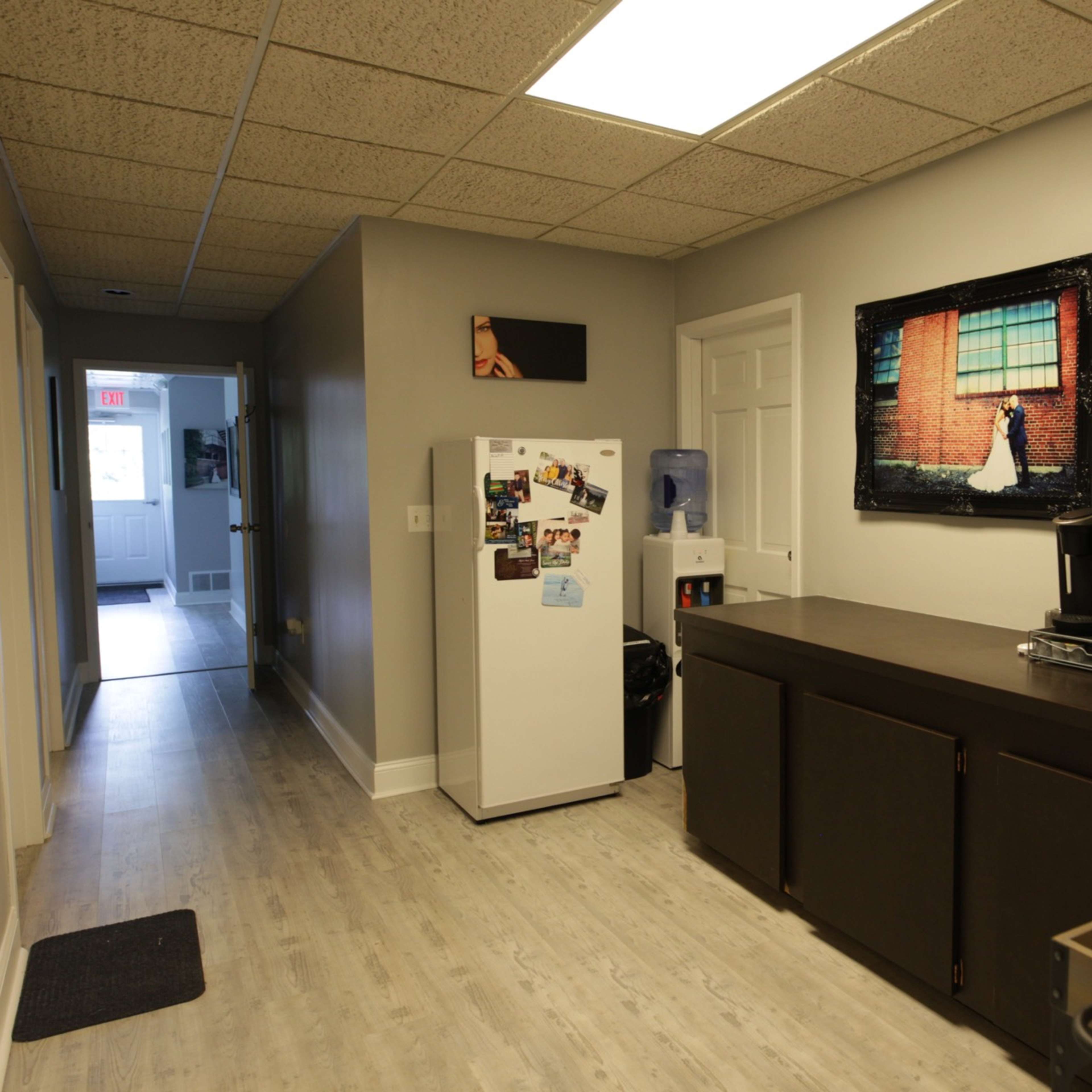 A narrow kitchen features a dark countertop, a white refrigerator, and a wall-mounted photo, with a hallway leading to a door at the far end.
