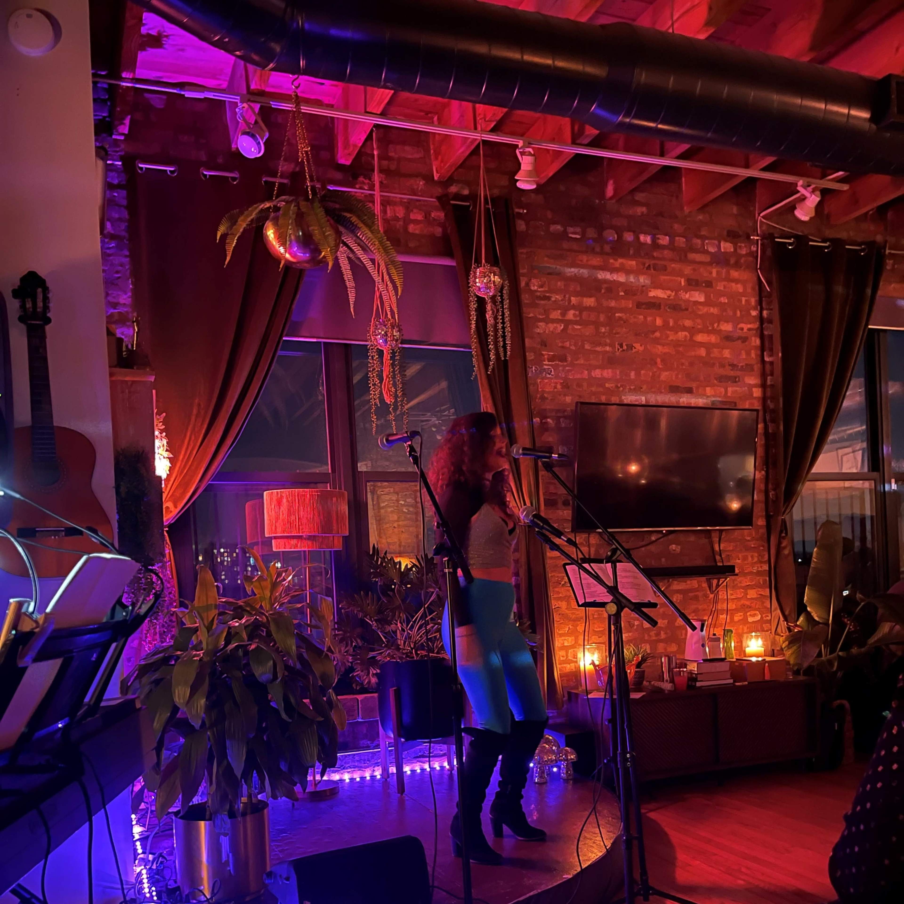 A performer stands on a stage in a dimly lit venue, surrounded by plants and decorative lights, while microphones and musical instruments are set up nearby.