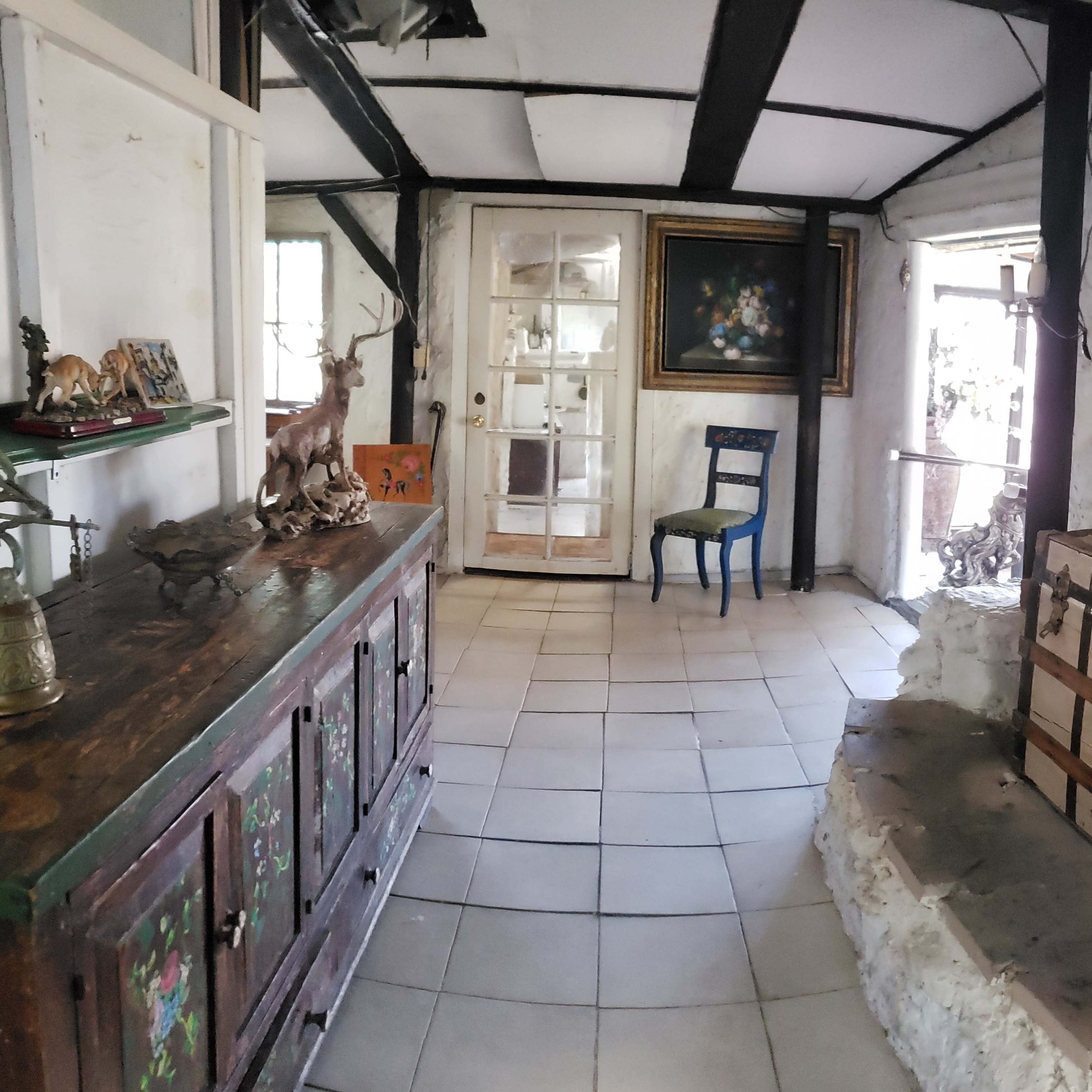 A rustic room featuring a wooden cabinet, a blue chair, a large painting, and a chest in a stone-walled space with a door leading outside.