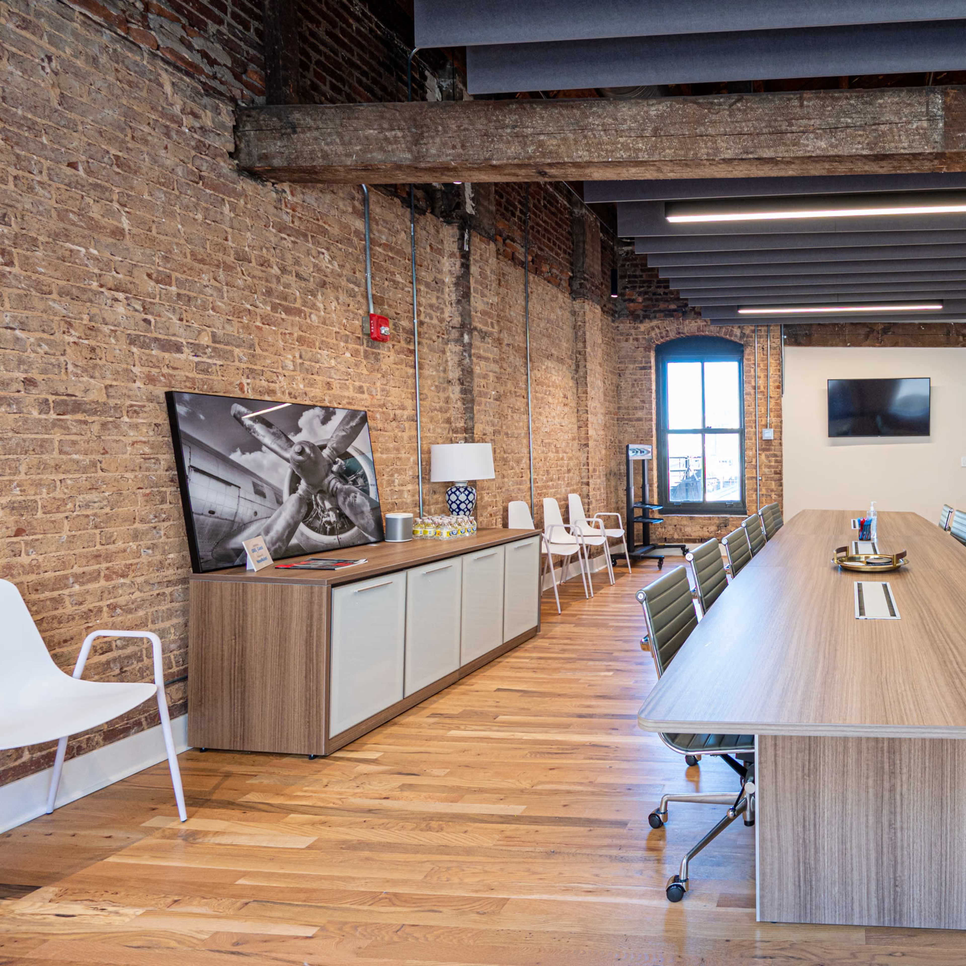 A modern conference room with a long wooden table, white chairs, exposed brick walls, and large windows.