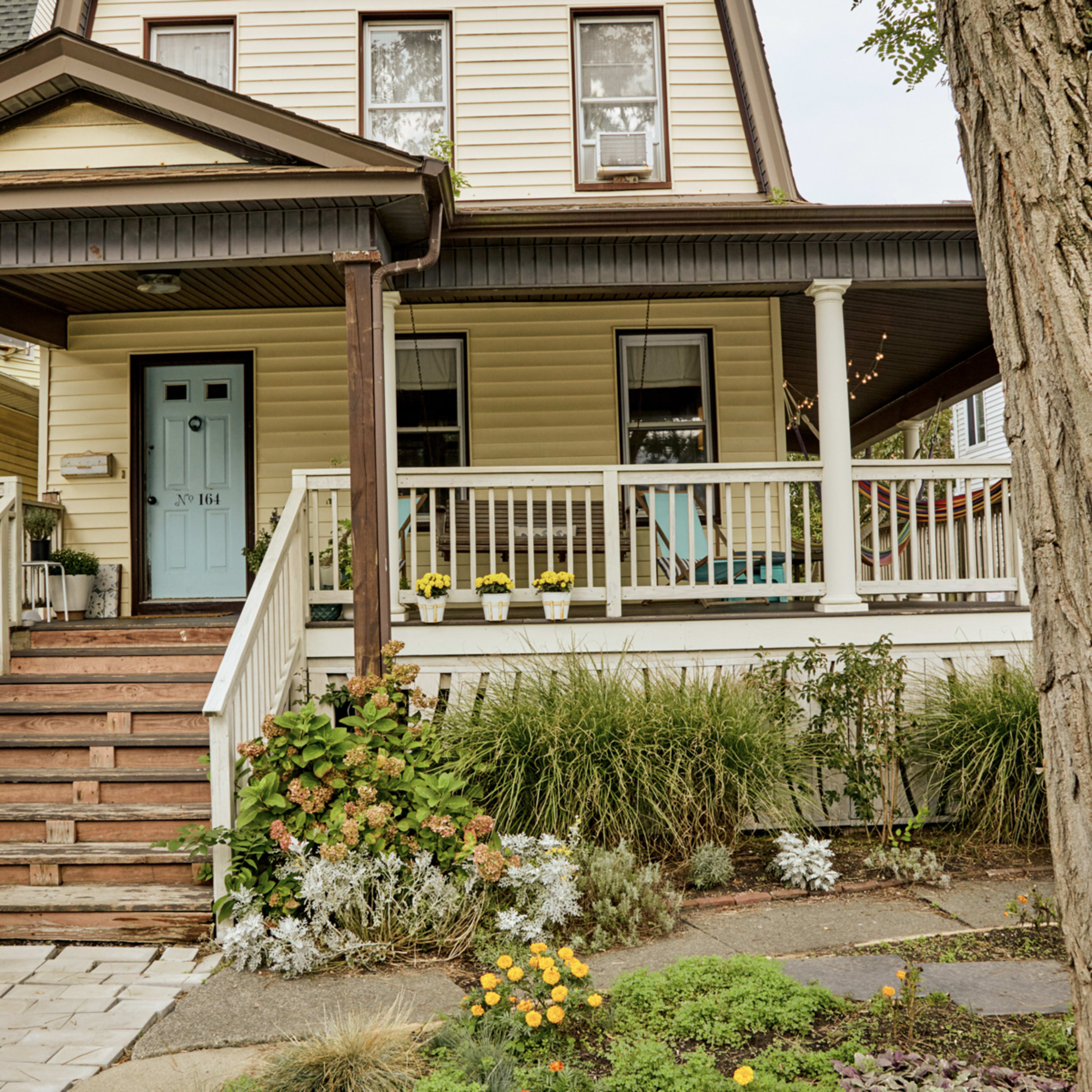 A two-story house with a front porch, wooden stairs, a blue door, and a garden filled with flowering plants.