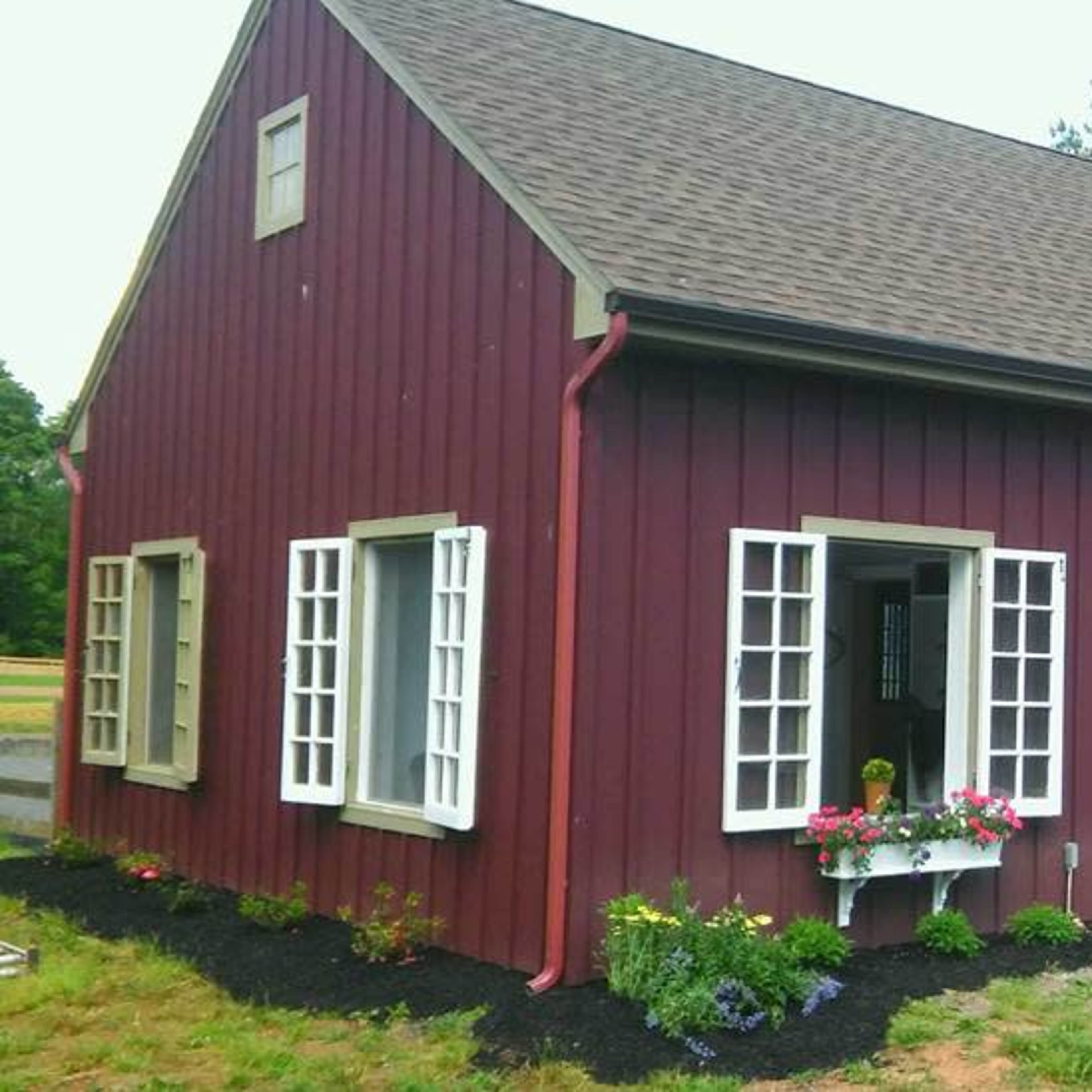 A red wooden house with a steep roof features large arched double doors and multiple windows, surrounded by flower boxes and gravel landscaping.