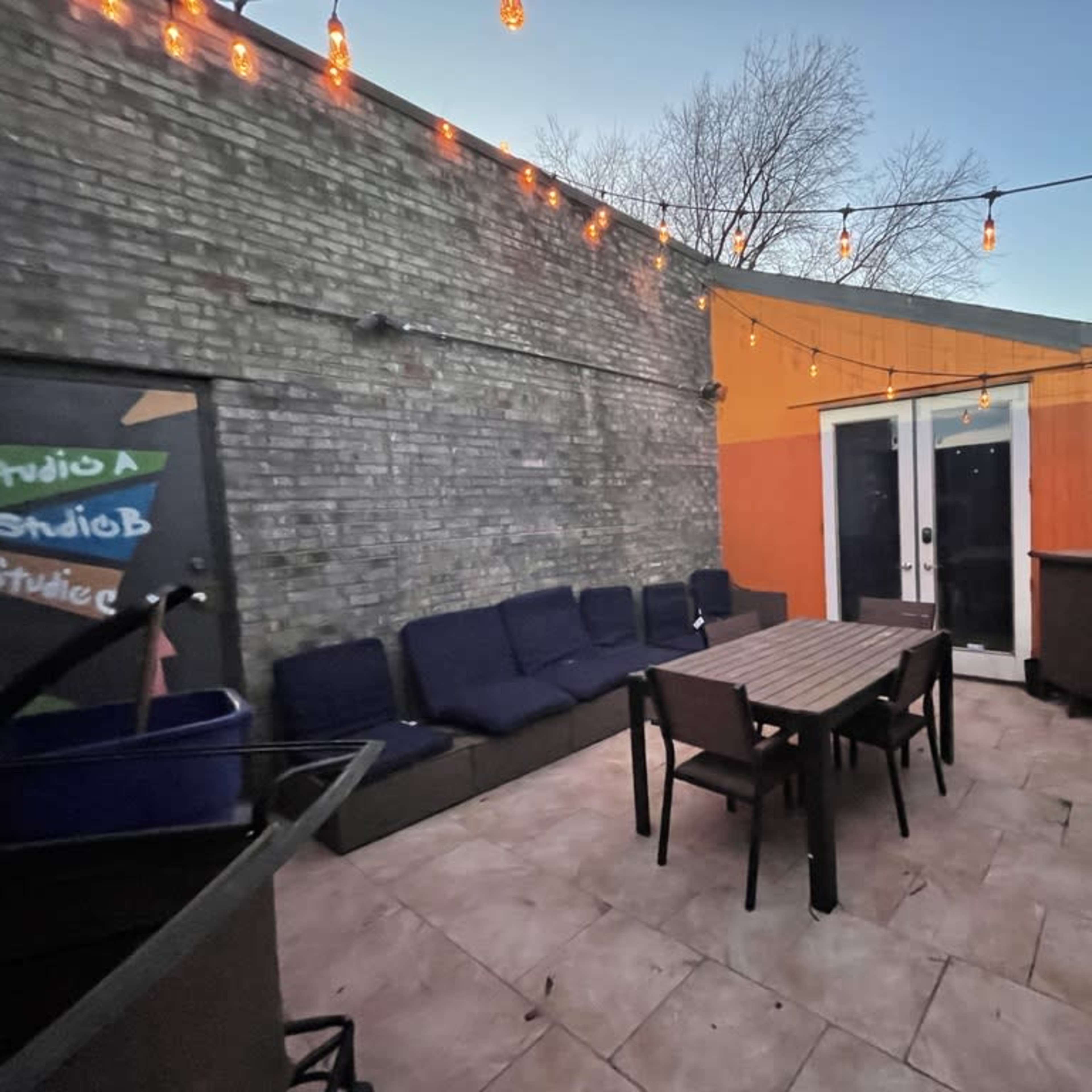 An outdoor patio space with a large table surrounded by chairs, a seating area with a couch, and string lights overhead.