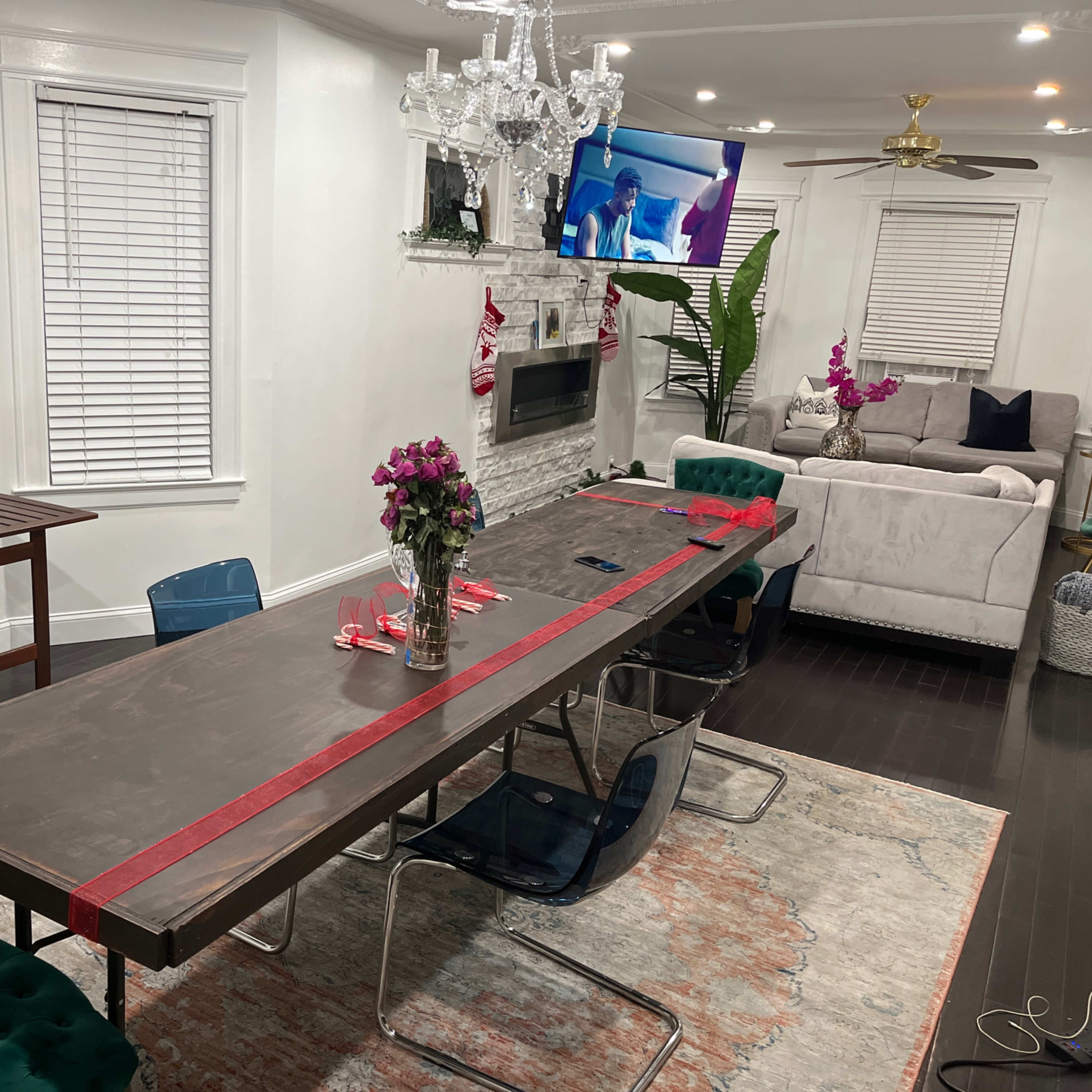 A long dining table is set with red ribbon and flowers, surrounded by chairs, in a room featuring a television, a sofa, and decorative plants.