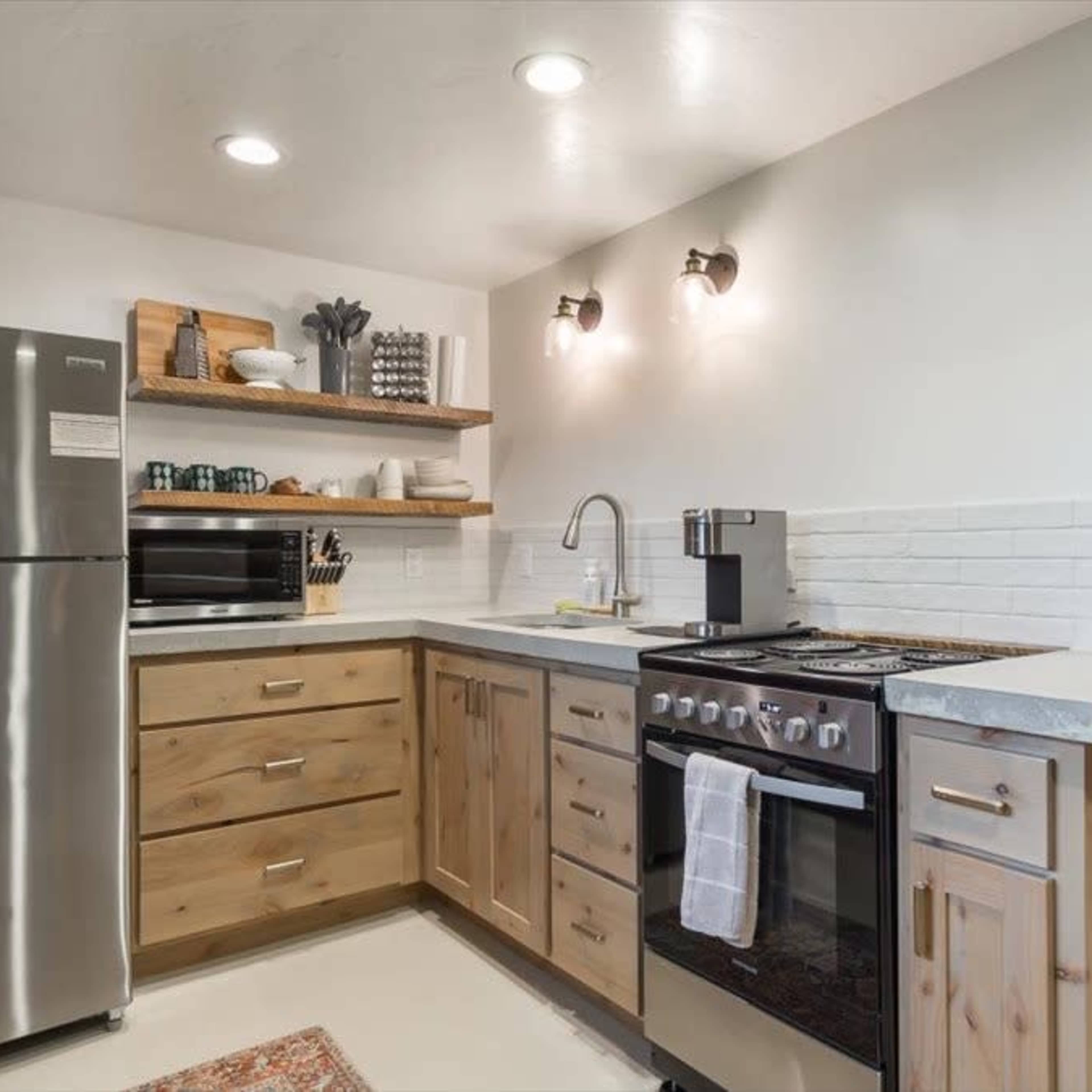 A modern kitchen with wooden cabinets, a stainless steel refrigerator, a stove, a microwave, and wall-mounted lights.