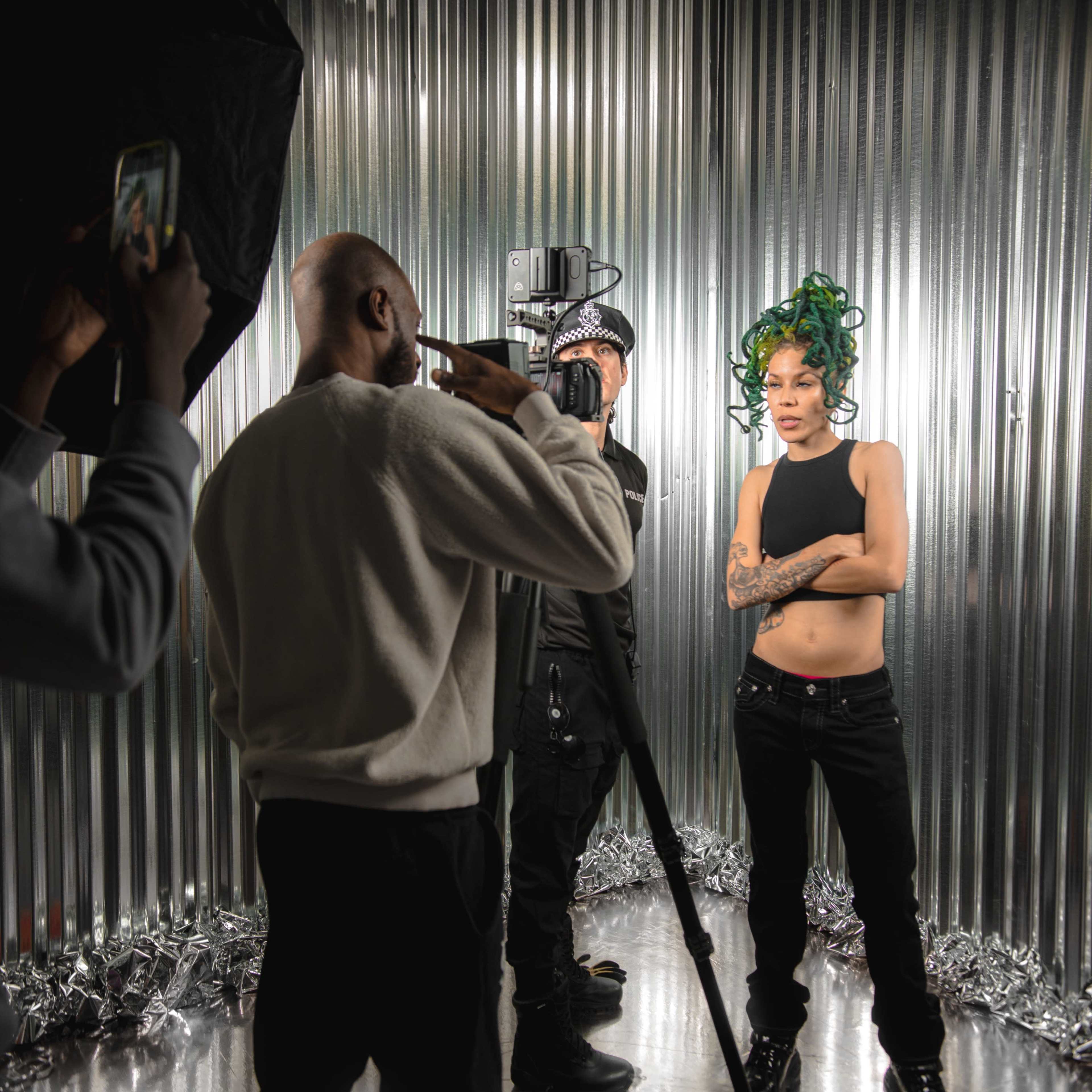 A model stands with crossed arms in front of a camera setup, while a photographer adjusts the equipment in a studio with metallic walls.