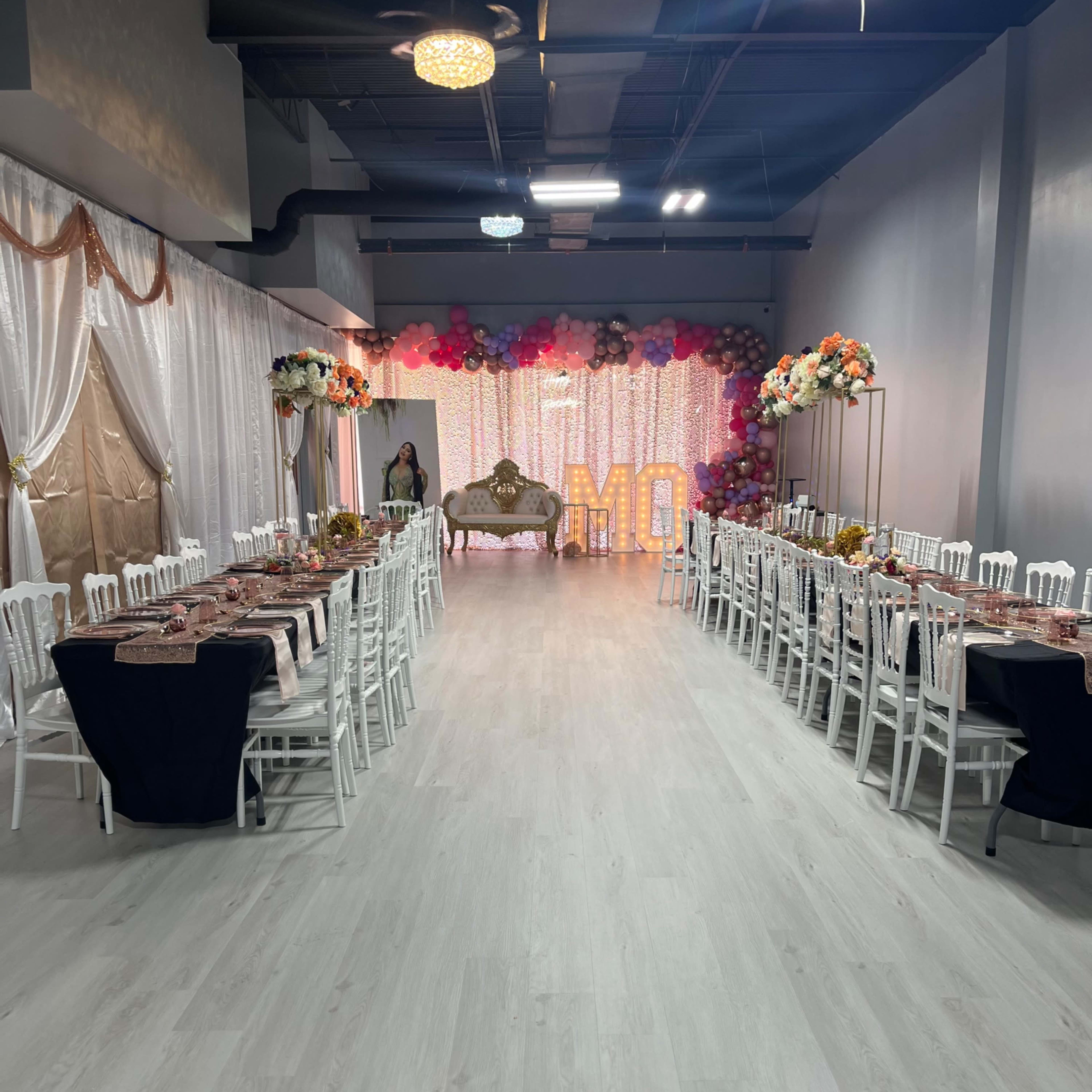 A spacious event venue set up with long tables and white chairs, decorated with floral arrangements and a sparkling backdrop.