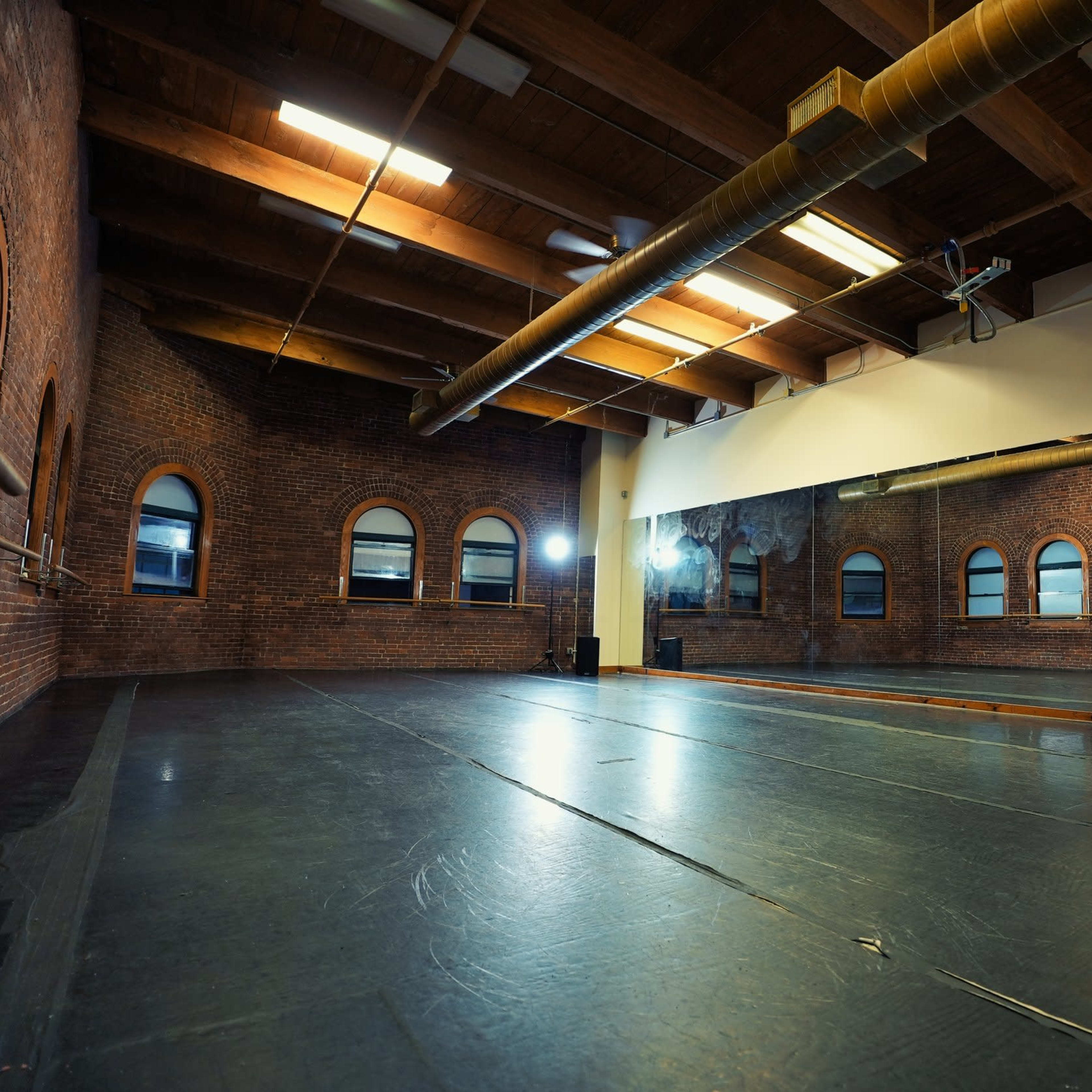 A spacious dance studio with brick walls, large windows, and a wooden ceiling.