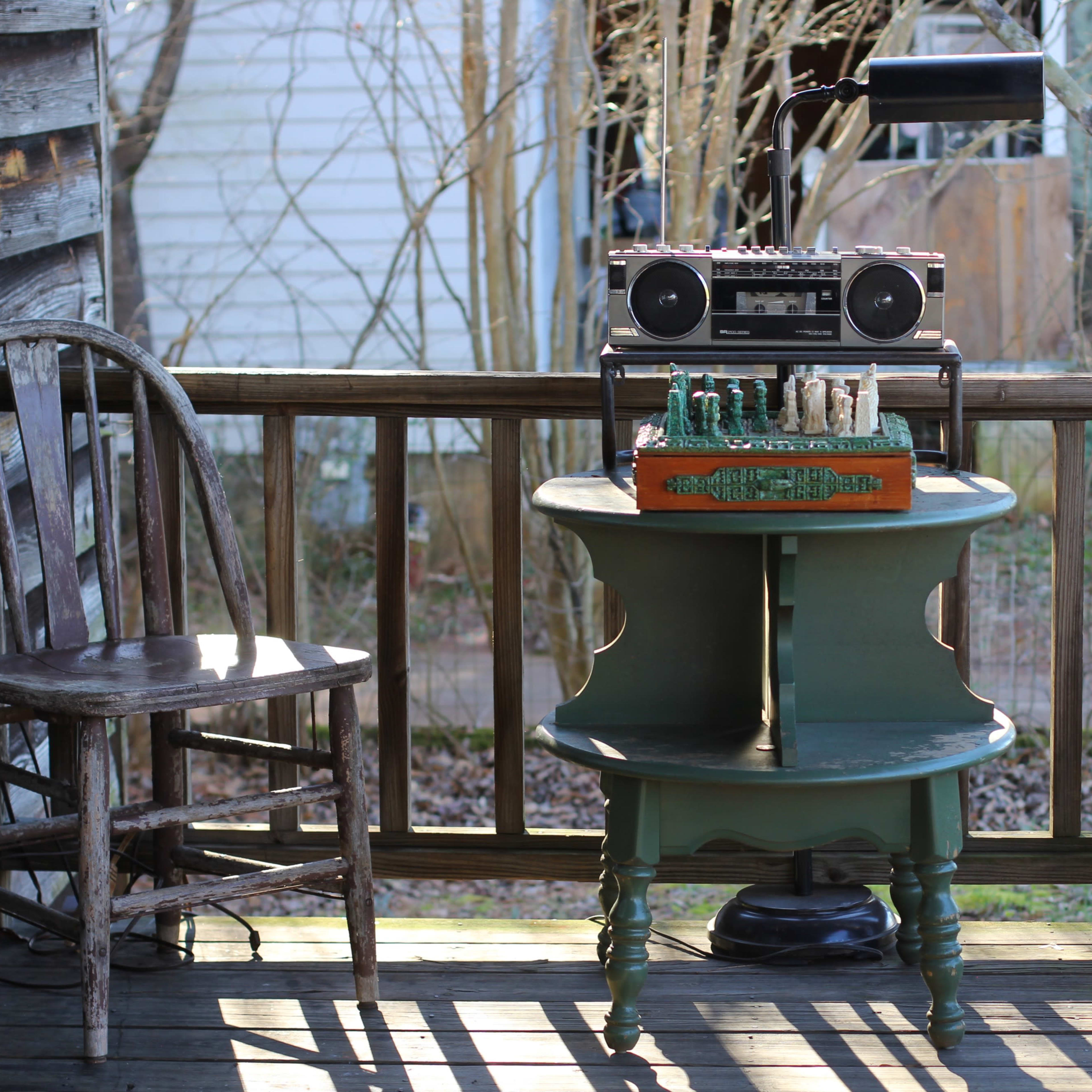 An old wooden porch features a round table with a vintage boombox and a decorative box, flanked by two mismatched chairs.