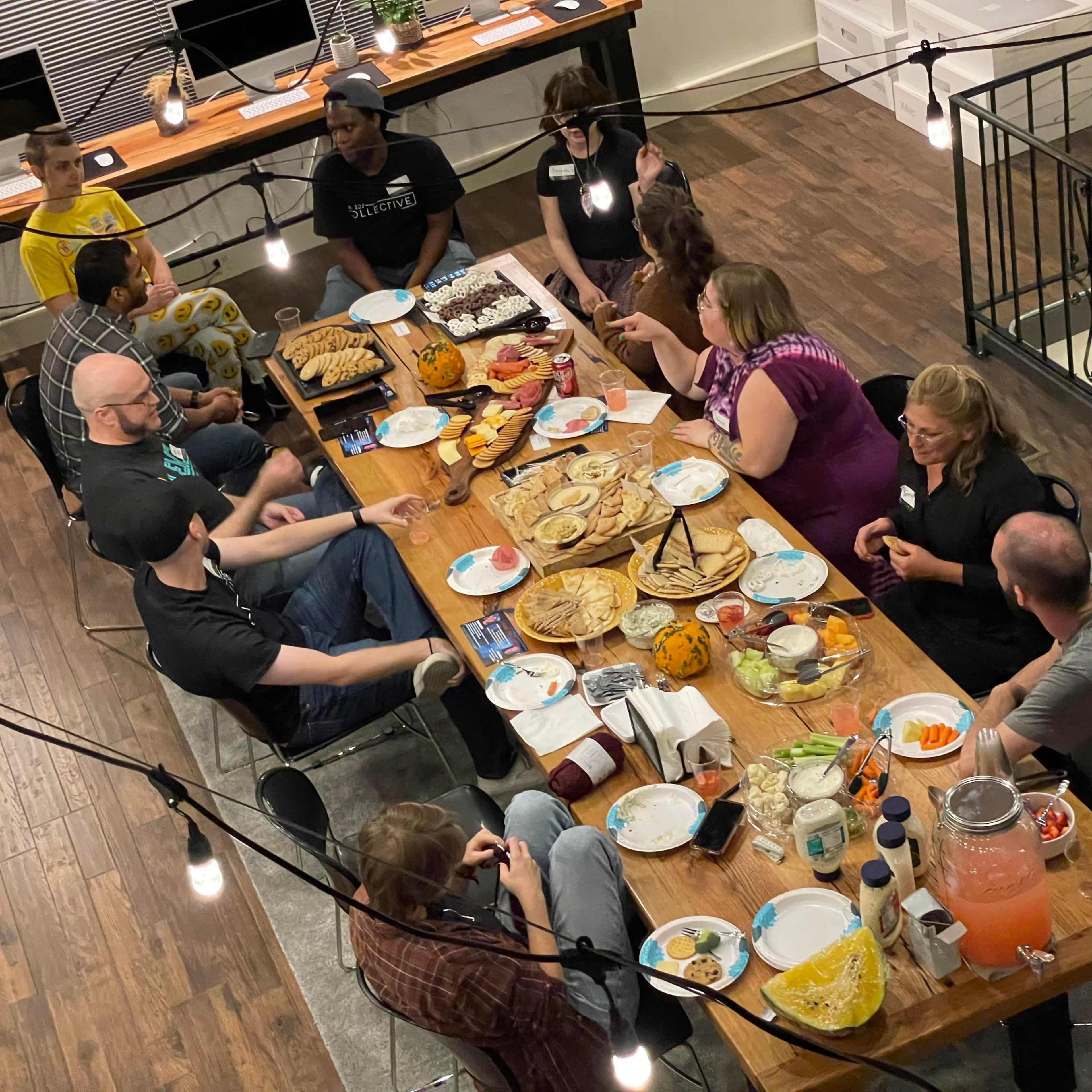 A group of people gathers around a large table filled with various food items, engaged in conversation under overhead string lights.