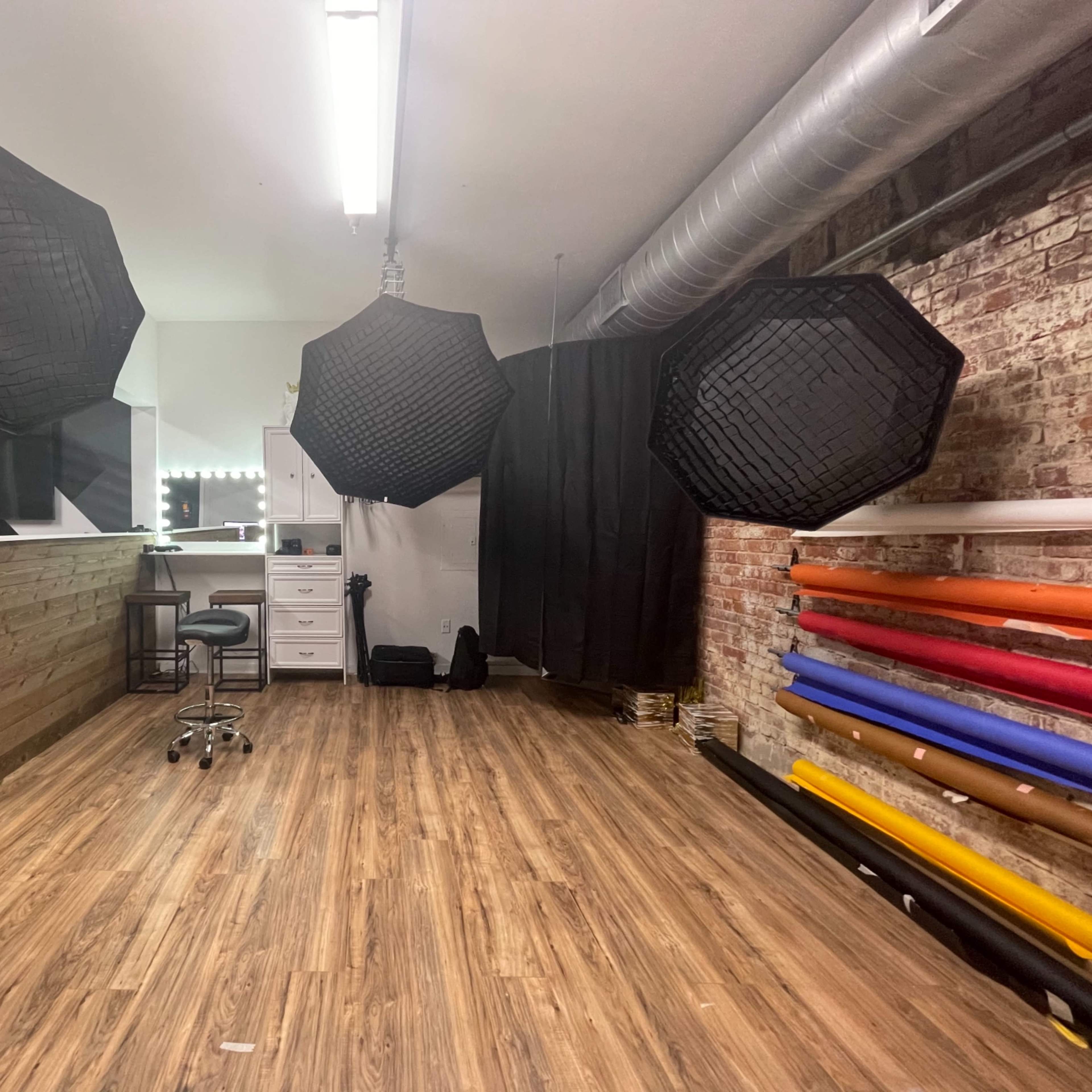 A photography studio with three large softboxes hanging overhead, a wooden reception desk, a chair, and rolls of colored backdrops along the wall.