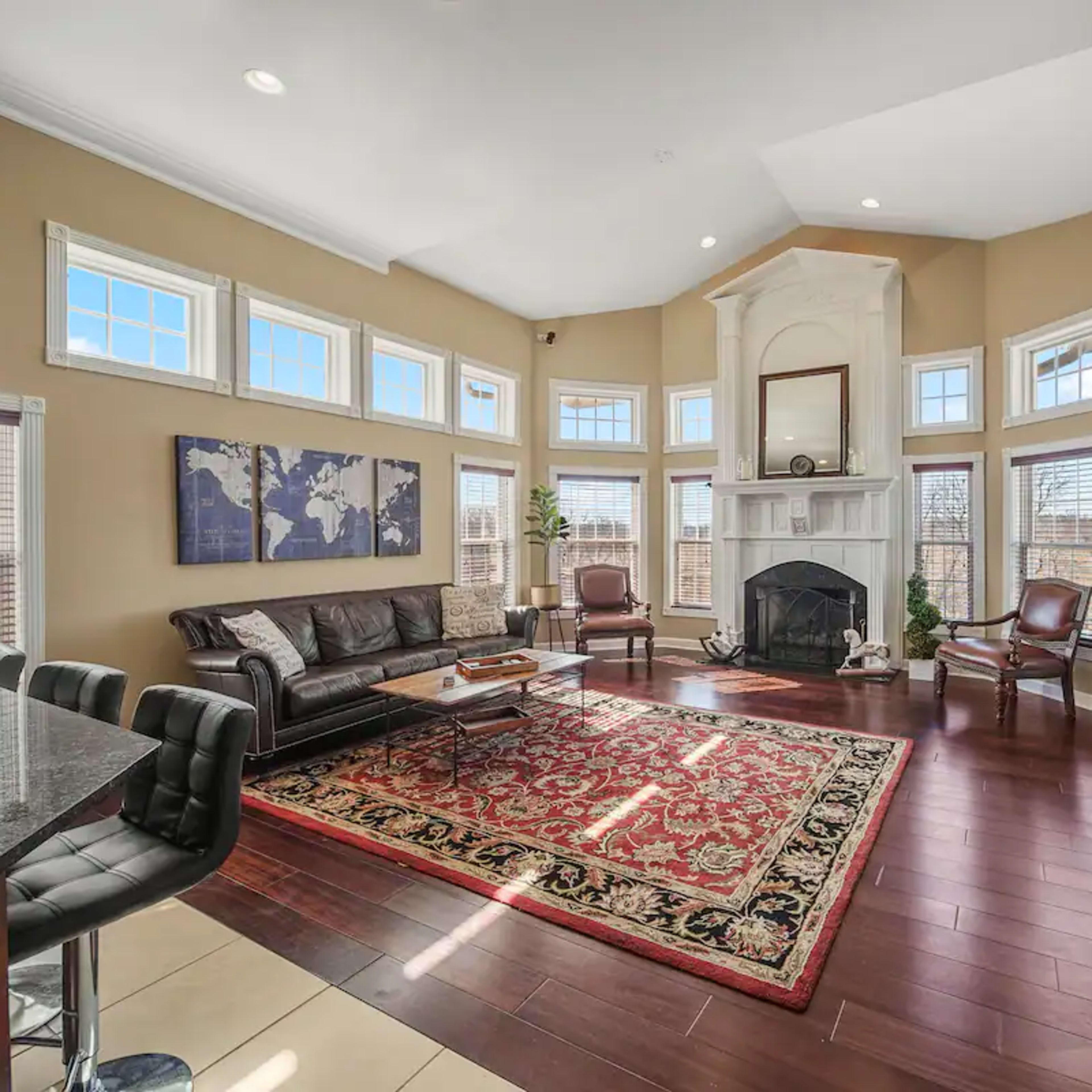 A spacious living room features a large bay window, a fireplace, and a patterned area rug, with seating arranged around a coffee table.
