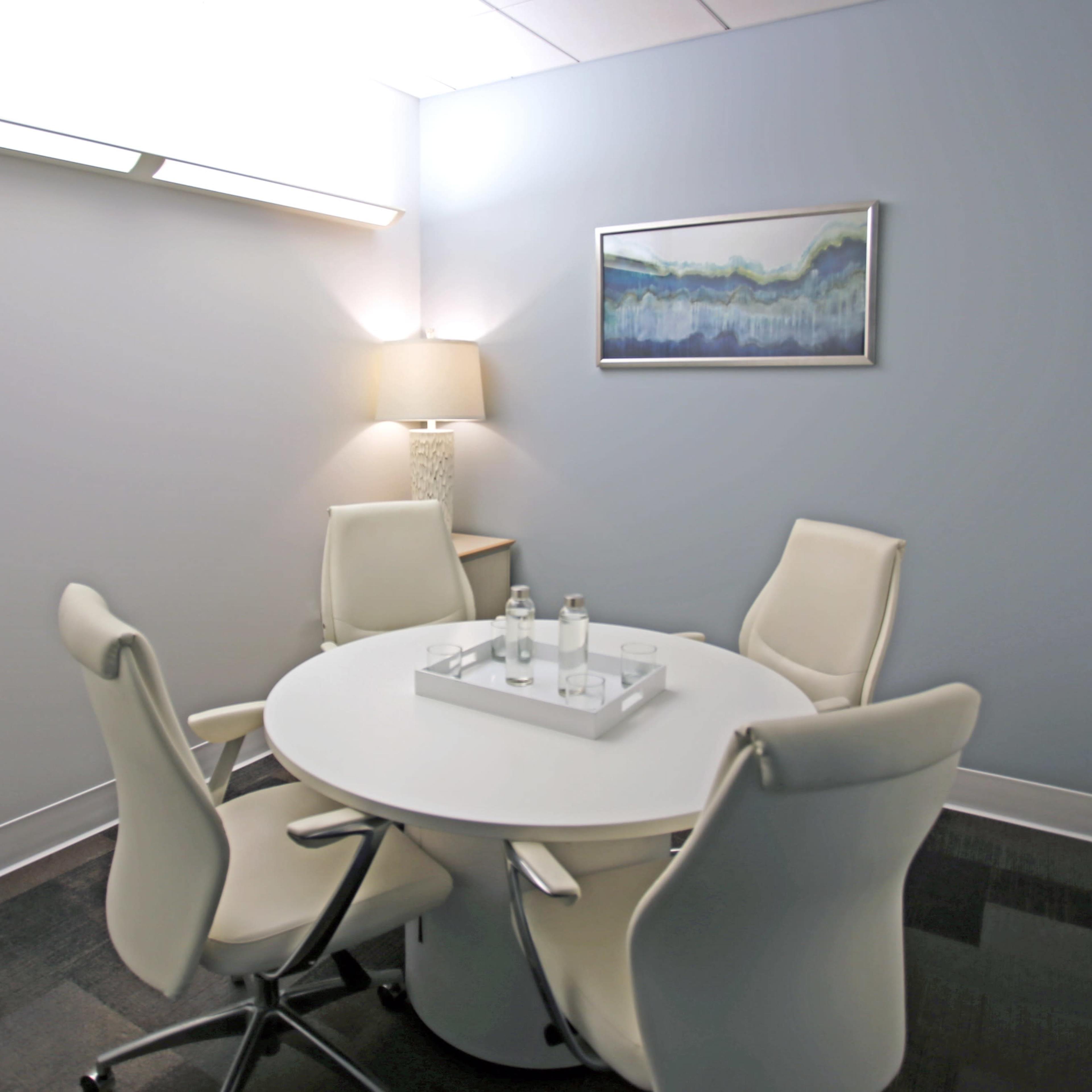 A small meeting room features a round table surrounded by four white chairs, with a lamp and a framed artwork on the walls.