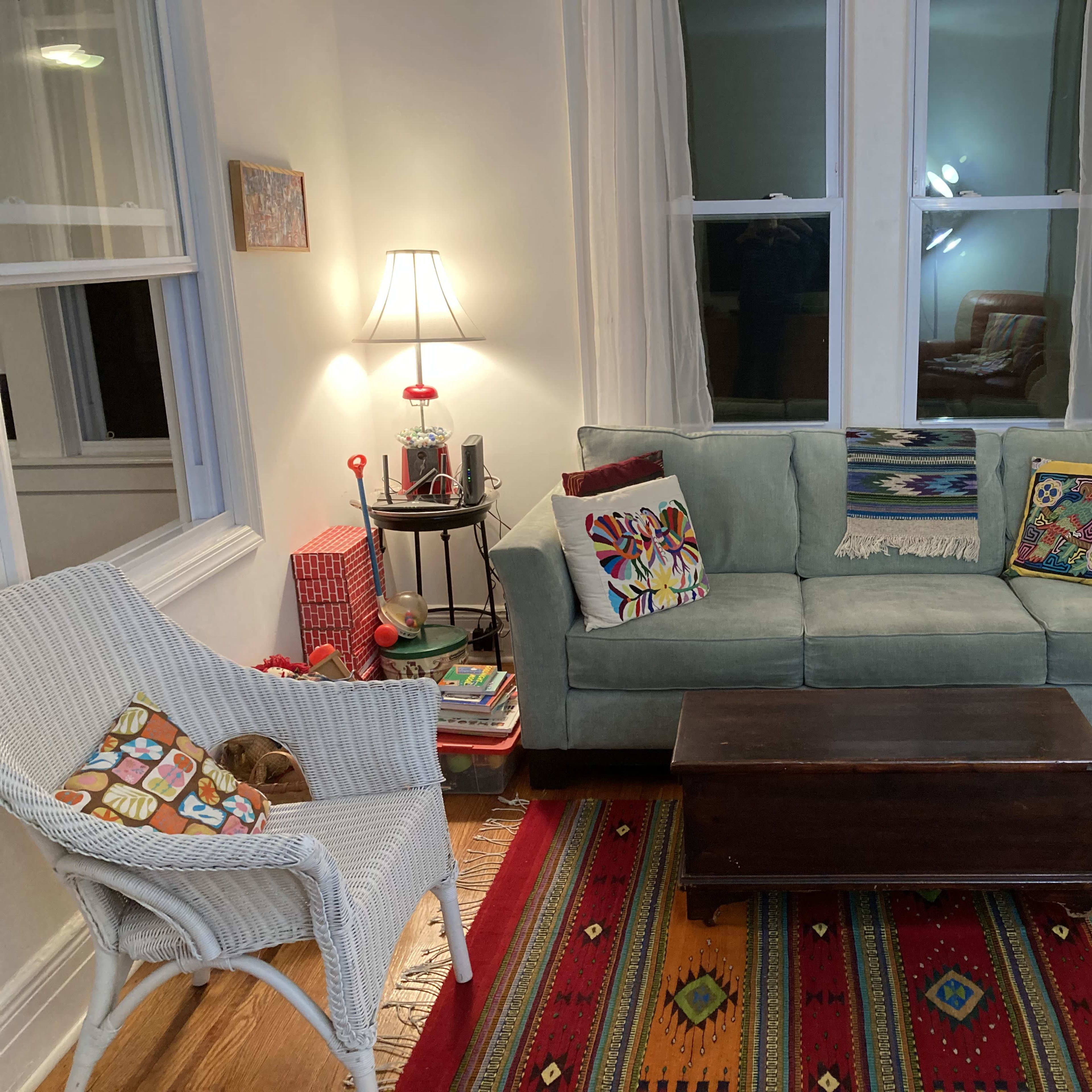 The living room features a green couch with decorative pillows, a wooden coffee table, and a white wicker chair on a patterned rug, illuminated by two lamps.