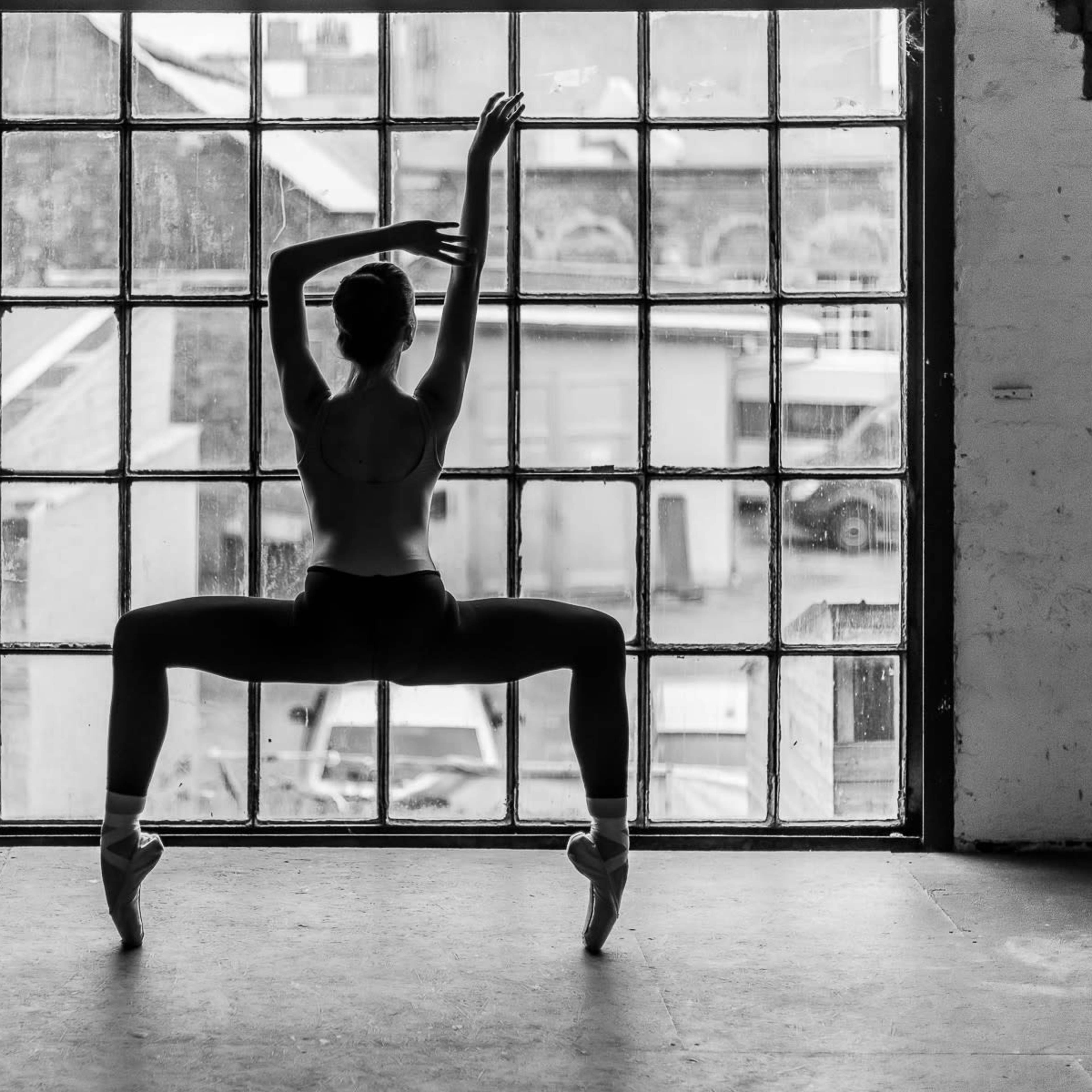 A dancer stands in a wide stance with arms raised, silhouetted against a large paneled window.